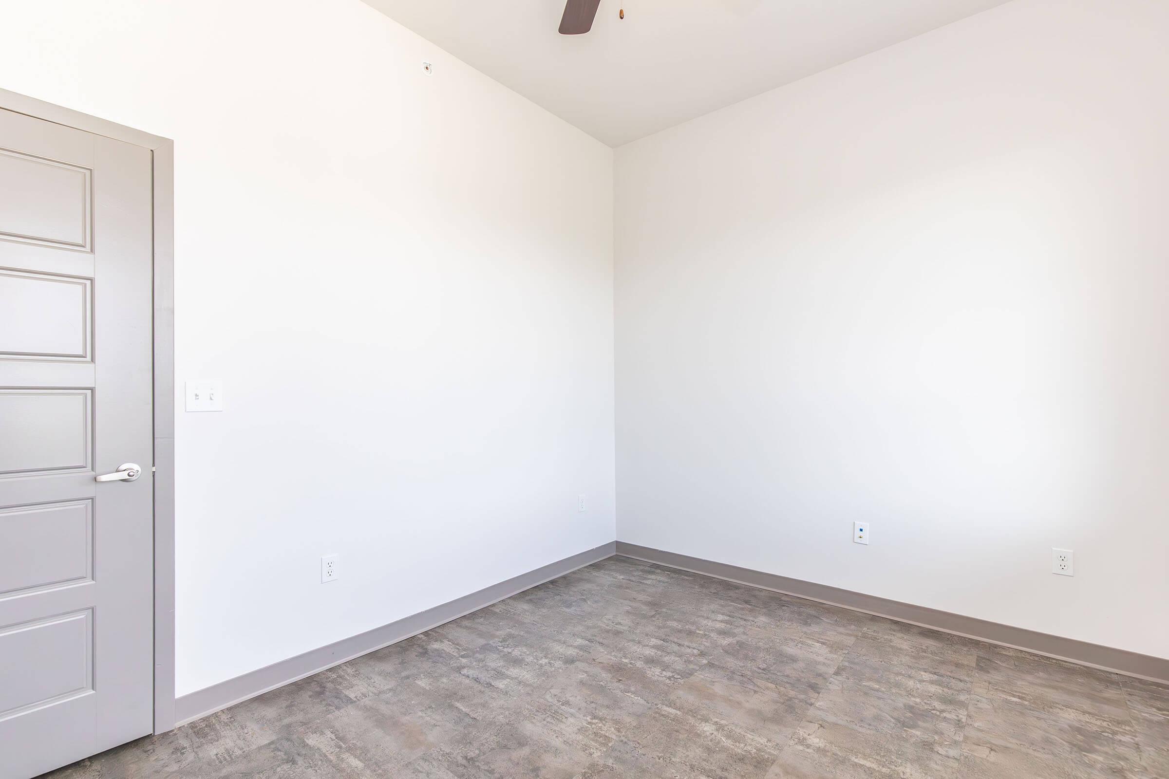 A spacious, empty room with light-colored walls, featuring a gray door on the left and a smooth floor. The room has a ceiling fan and ample natural light, creating a bright and airy atmosphere. Ideal for customization or use as a living space, office, or studio.