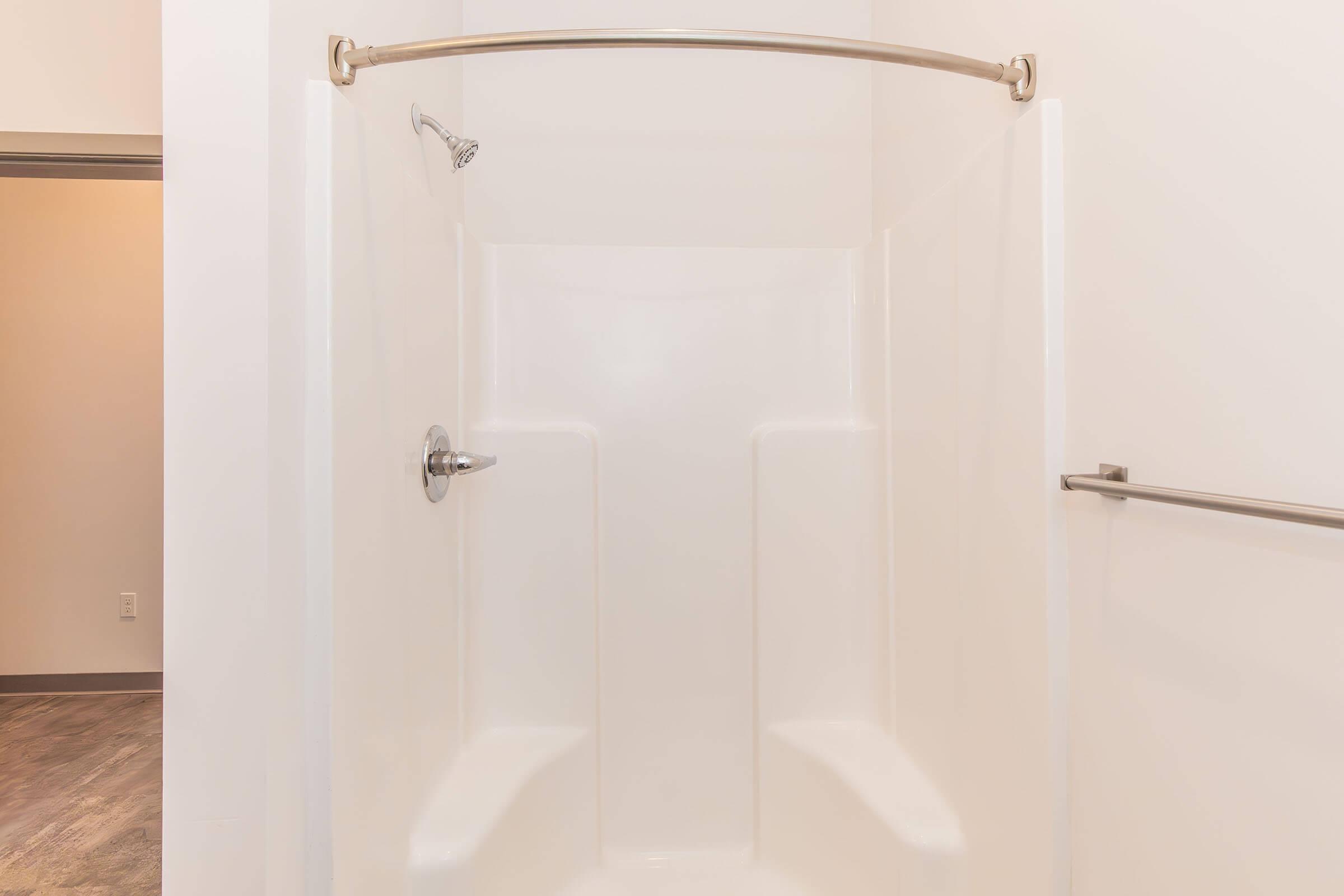 A clean, modern shower stall with a white interior, featuring a curved shower rod and a handheld showerhead. The backdrop is minimal, with light-colored walls and a glimpse of a doorway. The floor appears to have a wood-like finish.