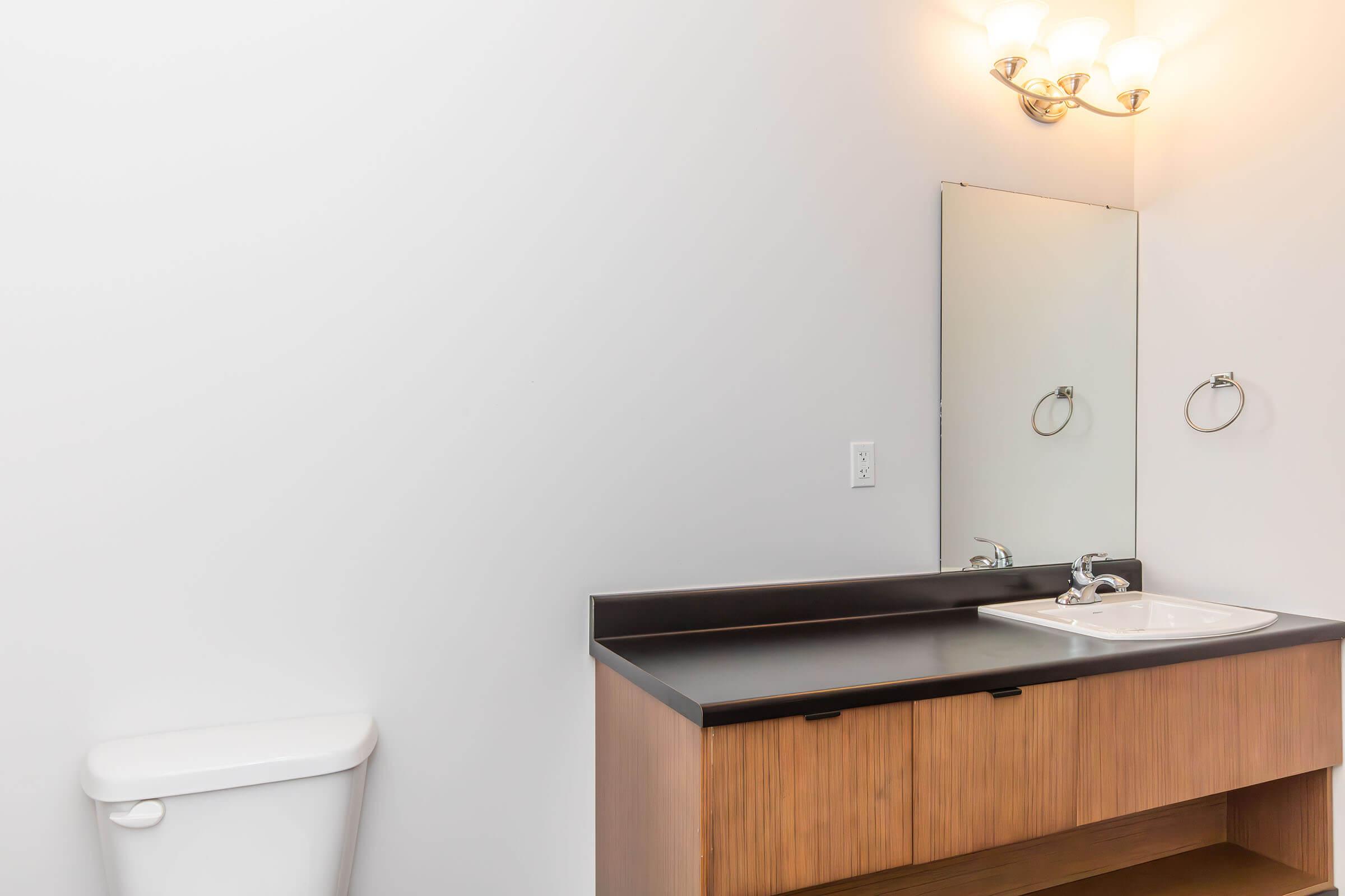 A modern bathroom featuring a white toilet, a wood vanity with a black countertop, and a sink. A mirror is mounted above the sink, and there is a wall-mounted towel ring. Soft lighting is provided by a fixture with three bulbs above the mirror, against a plain white wall.