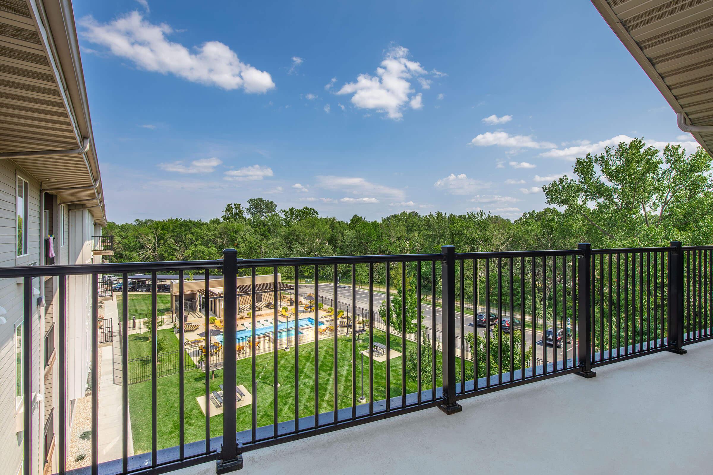 A balcony view overlooking a landscaped area with a swimming pool and lounge chairs. The sky is bright blue with a few clouds, and there are trees in the background. The scene captures a vibrant and inviting outdoor space.