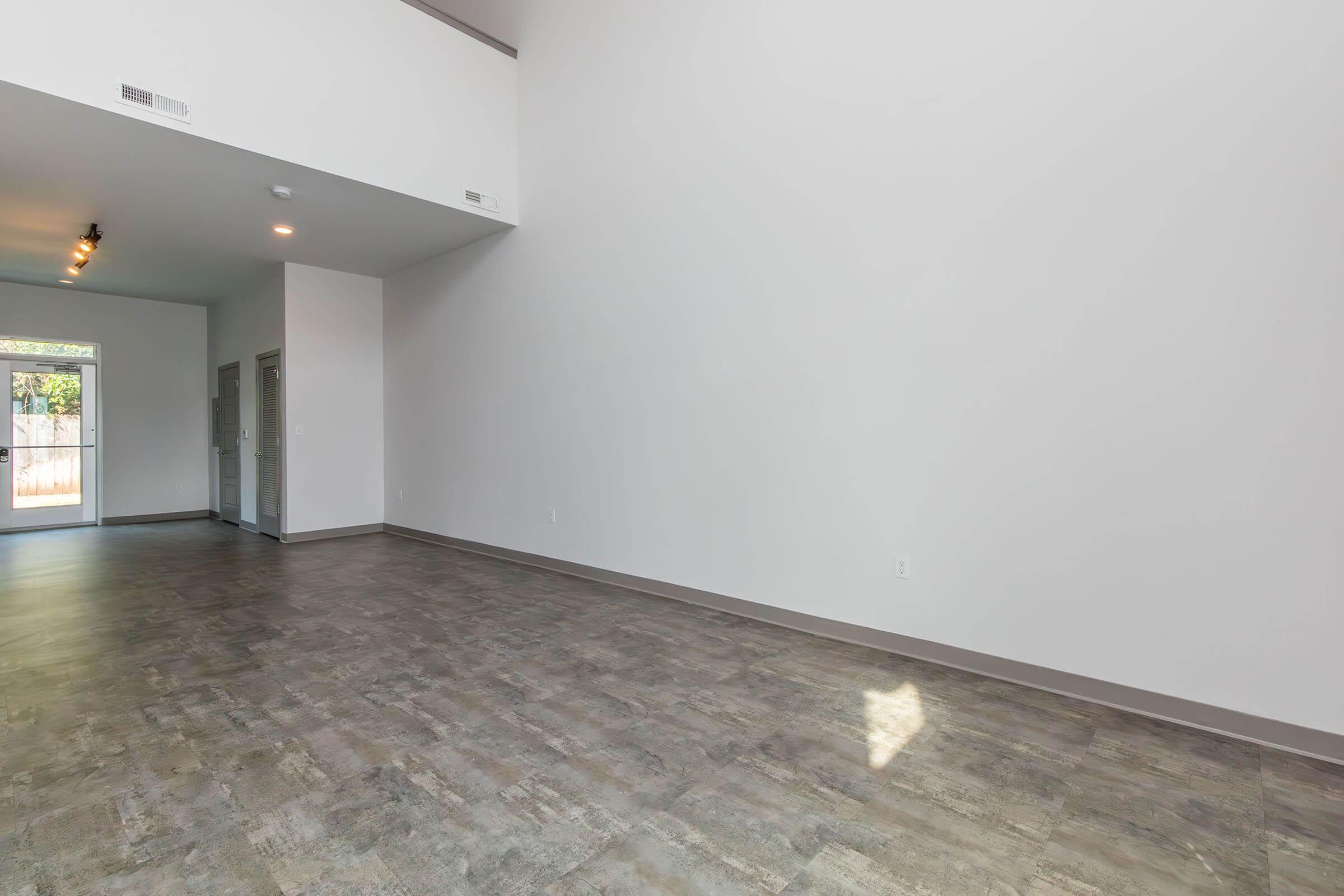A spacious, modern interior space with high ceilings and neutral-colored walls. The flooring is light-colored and has a smooth finish. Natural light enters through large windows, illuminating the area. The room is empty, creating a minimalist atmosphere with potential for various layouts and furnishings.