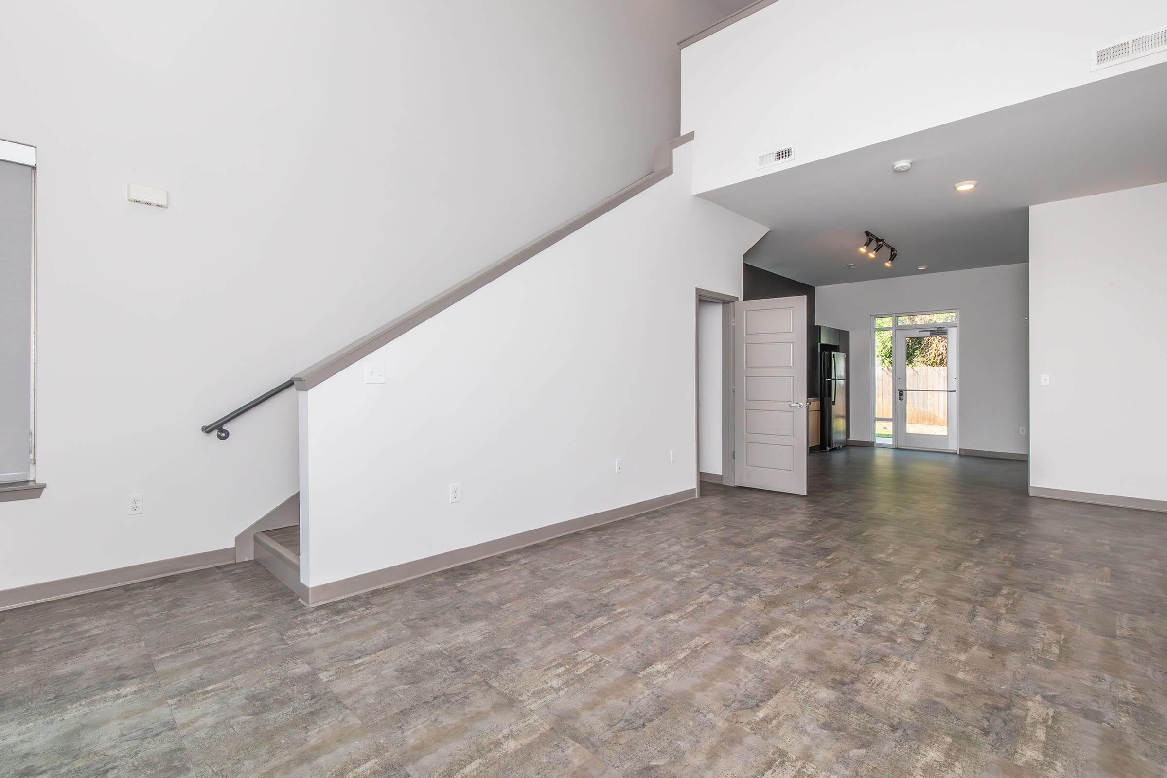 Spacious modern interior showing a living area with a staircase, gray walls, and a large window. The room features laminate flooring and an open layout leading to another room, partially visible with a door and faint natural light from an exterior entrance.