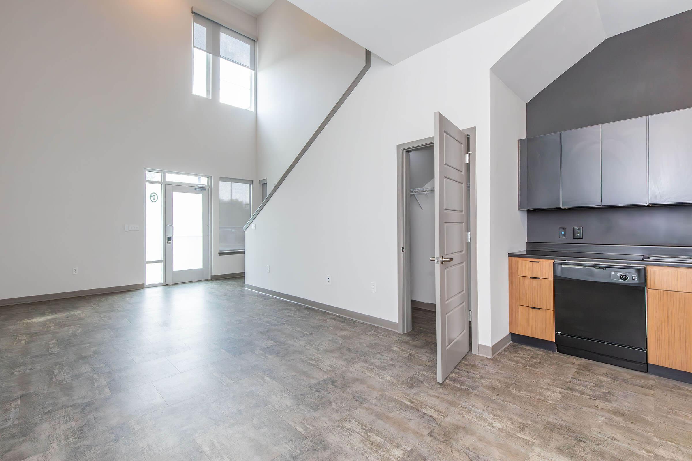 Modern interior of an apartment featuring an open layout with large windows, a staircase, and a kitchen area. The kitchen has black appliances and wooden cabinetry, while the living space has a neutral color scheme and tiled flooring. The front door is visible, leading to an entrance area.
