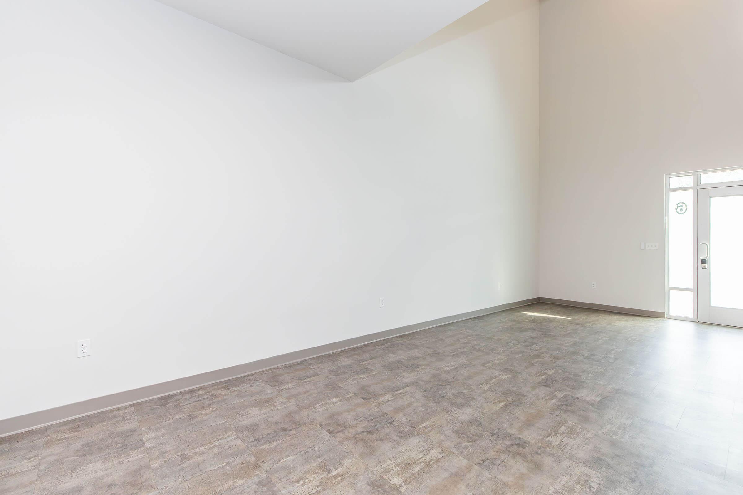 Spacious, empty room with a high ceiling and light-colored walls. A large window and door on one side allow natural light to enter, while the floor features a modern, neutral-toned surface. The space is ideal for various uses, such as a living area or office.