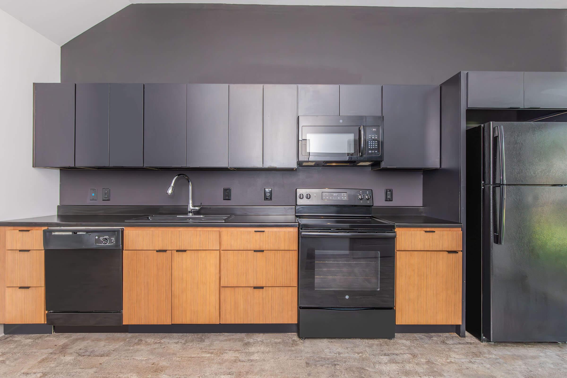 Modern kitchen featuring dark cabinets, a black countertop, stainless steel dishwasher, microwave, and oven. A sleek black refrigerator stands to the right, and the space has a minimalist design with wood accents. Natural light may be present through an unseen window, creating an inviting atmosphere.
