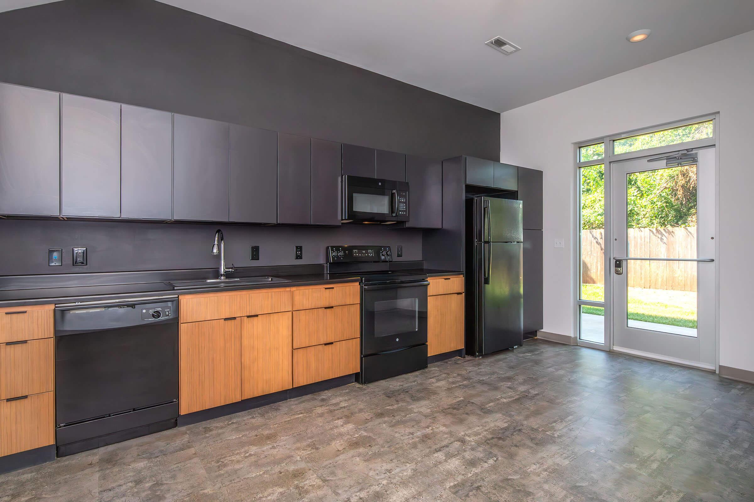 Modern kitchen featuring sleek black and wooden cabinetry, stainless steel appliances including a microwave and dishwasher, and a large window with glass doors leading to an outdoor space. The flooring is a neutral-toned material, complementing the contemporary design.