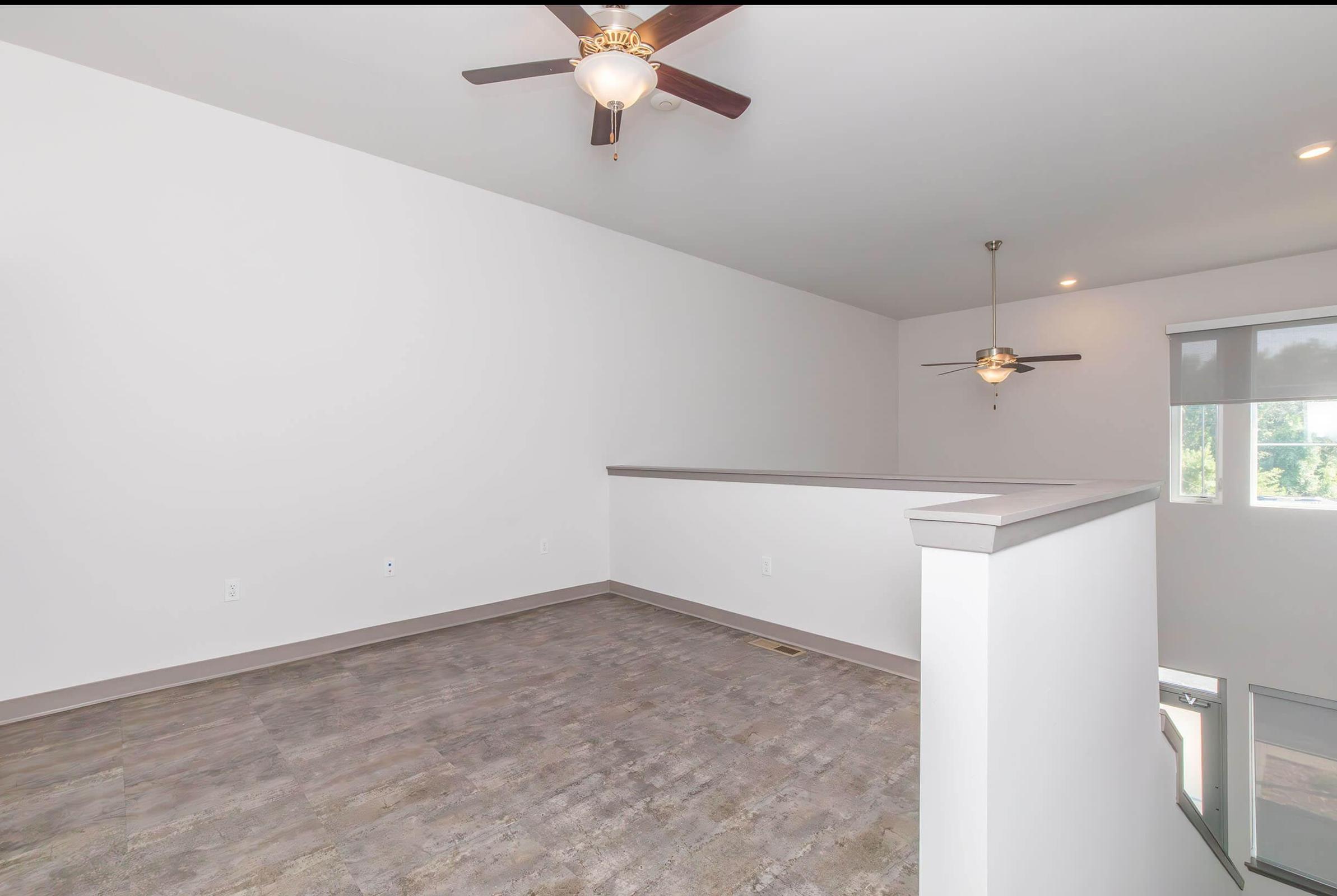 A spacious, empty room with light gray walls and a ceiling fan. The floor is covered in light-colored tiles. A railing indicates a upper-level view, and a large window allows natural light to enter. The layout is open and airy, suggesting potential for various uses or furnishings.