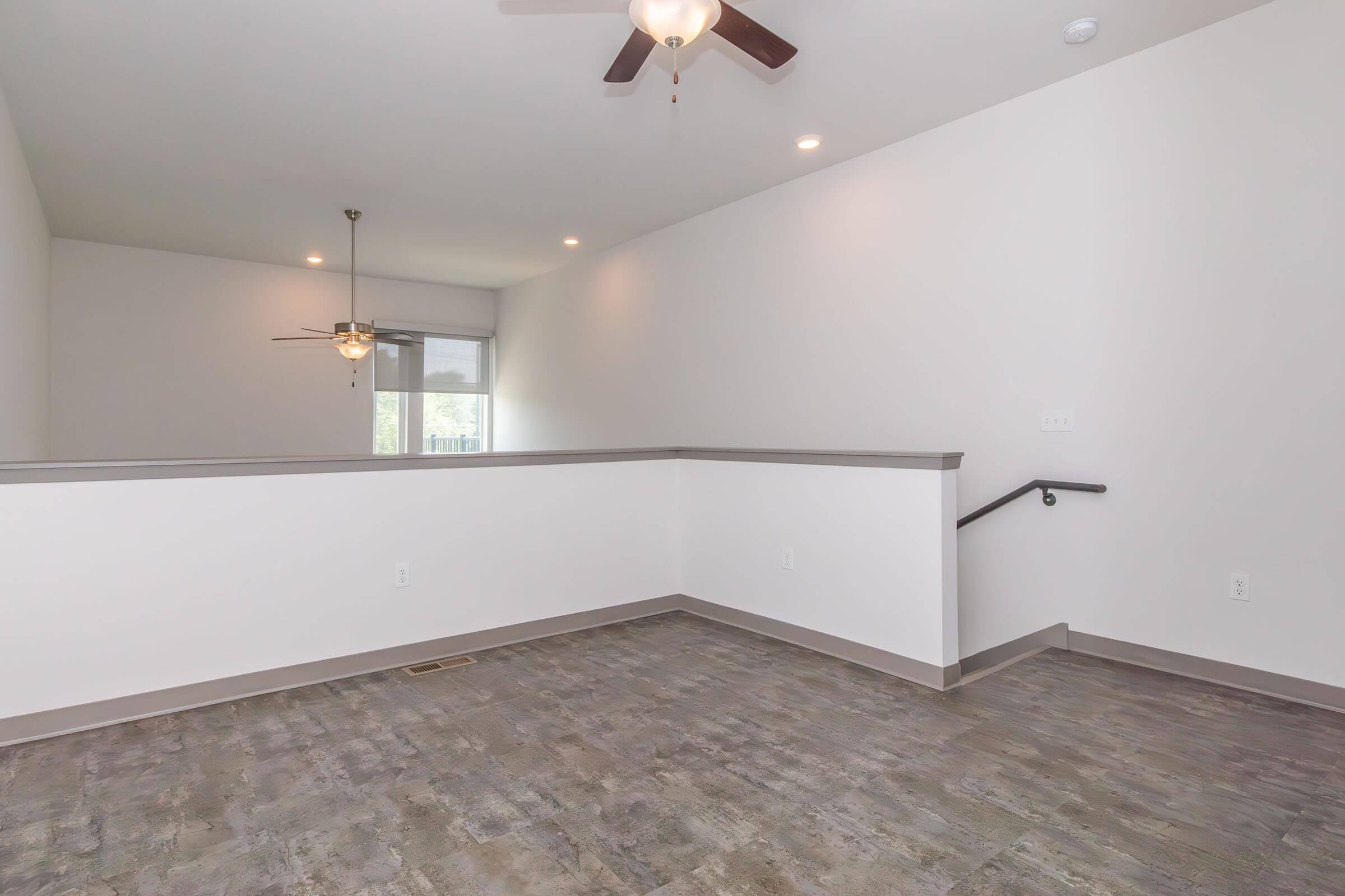 A spacious, empty room with light-colored walls, a ceiling fan, and large windows allowing natural light. The floor features a modern design with a wood-like appearance. A staircase leads to a different level, and the area is well-lit with overhead lights. Ideal for a living room or open-concept space.