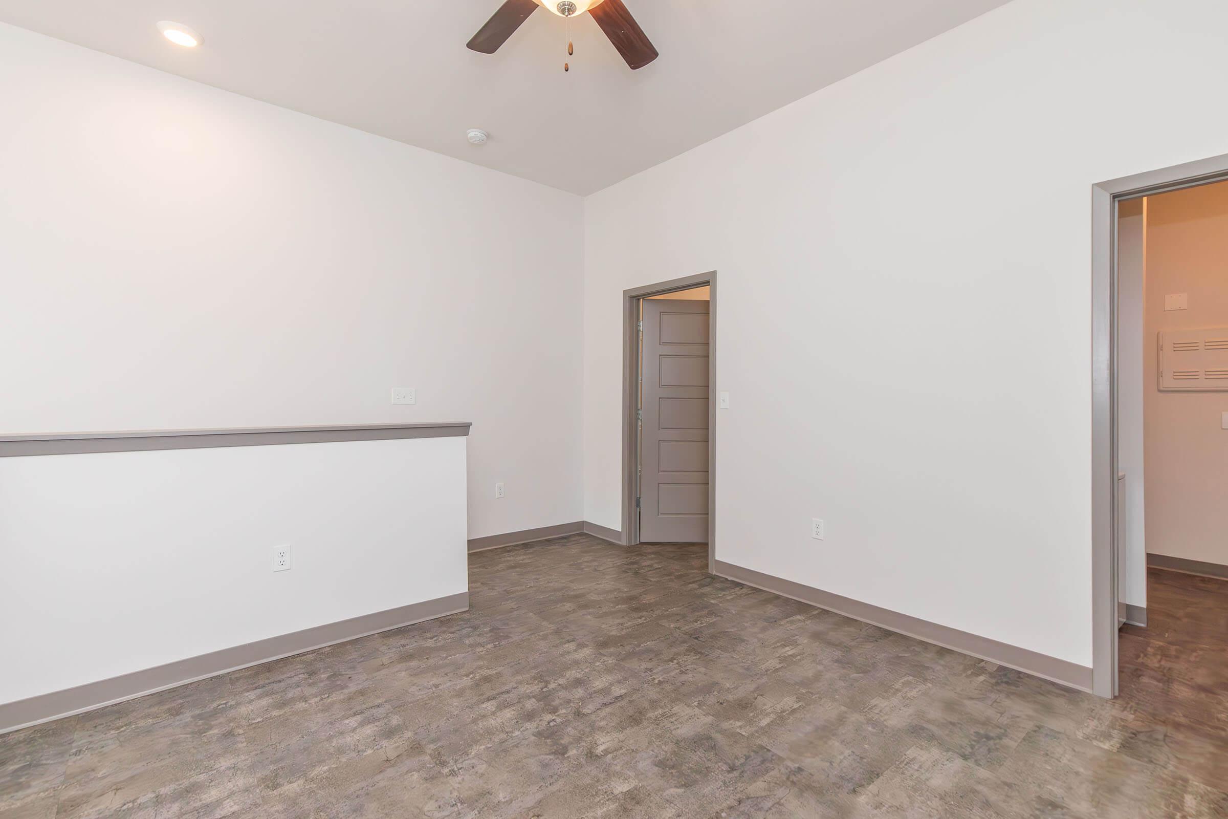 A spacious, modern room featuring light-colored walls, a ceiling fan, and a gray laminate floor. There is a partial wall or counter on the left, and an open doorway leading to another area on the right. The overall design is minimalist and bright, emphasizing simplicity and clean lines.