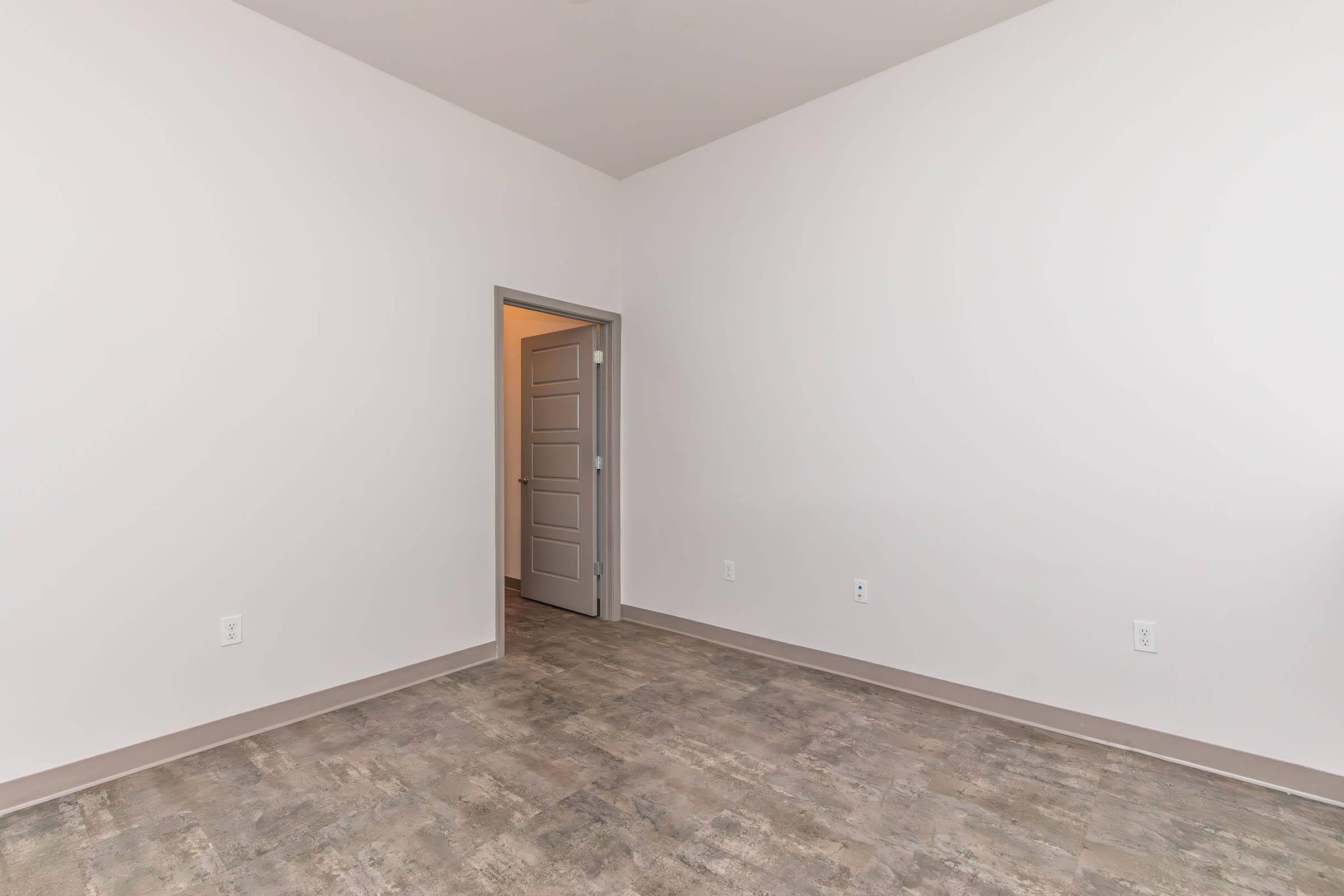A spacious, empty room with light-colored walls and a neutral-toned floor. A partially open door leads to another area, suggesting a potential living space. The room has no furniture or decorations, emphasizing its blank canvas for future design.