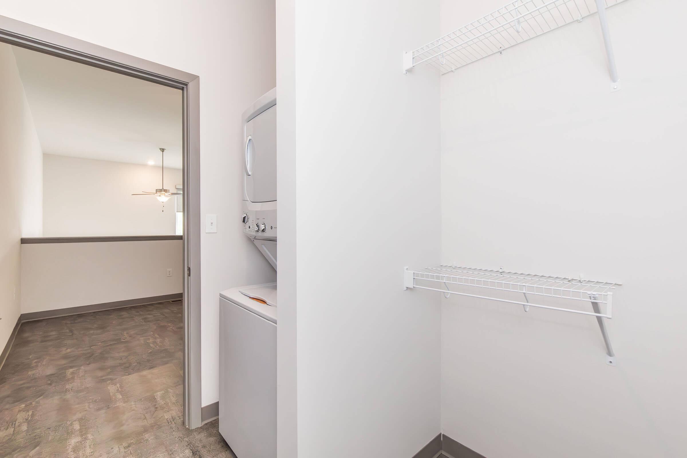 A laundry room featuring a stacked washer and dryer on the left, with a wire shelf above. The room has light-colored walls and a modern floor. In the background, a doorway leads to another room with dim lighting and a ceiling fan.