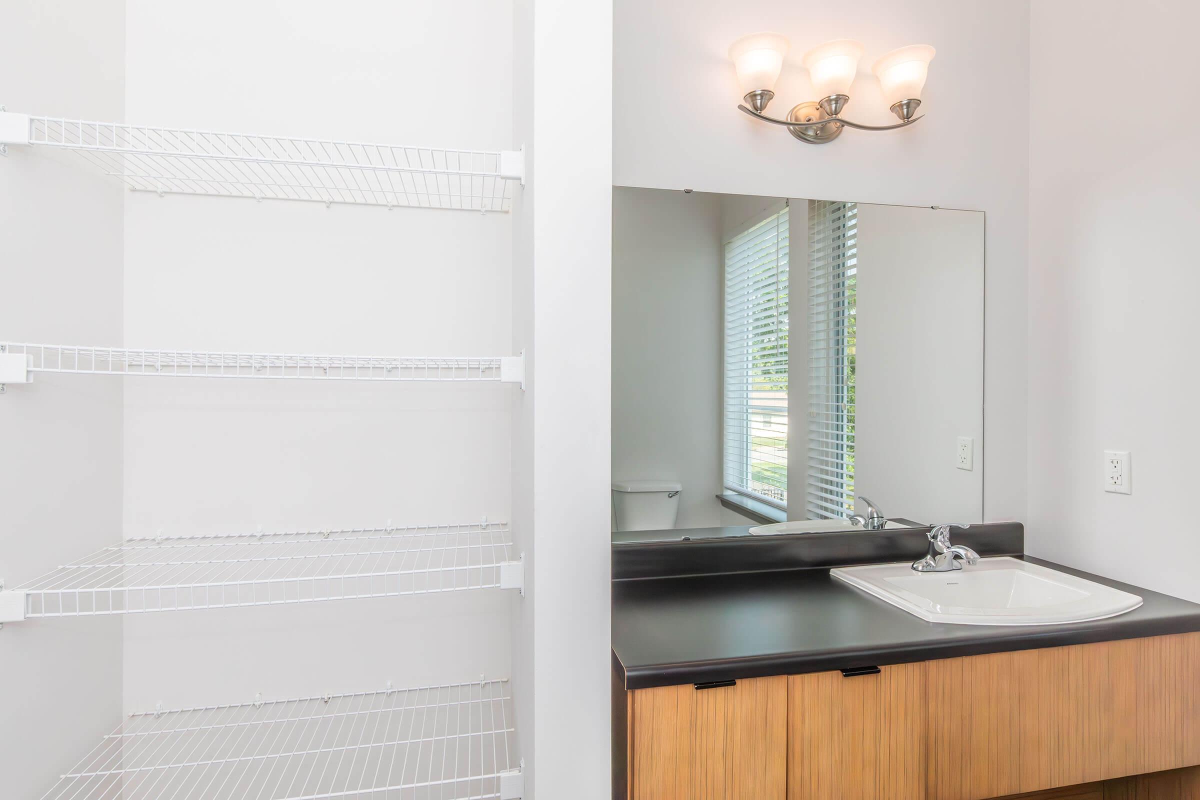 A bright and modern bathroom featuring a white shelf unit on the left, a sleek black countertop with a white sink, and a large wall mirror. Natural light flows in through a window with horizontal blinds, providing a clean and minimalistic aesthetic.