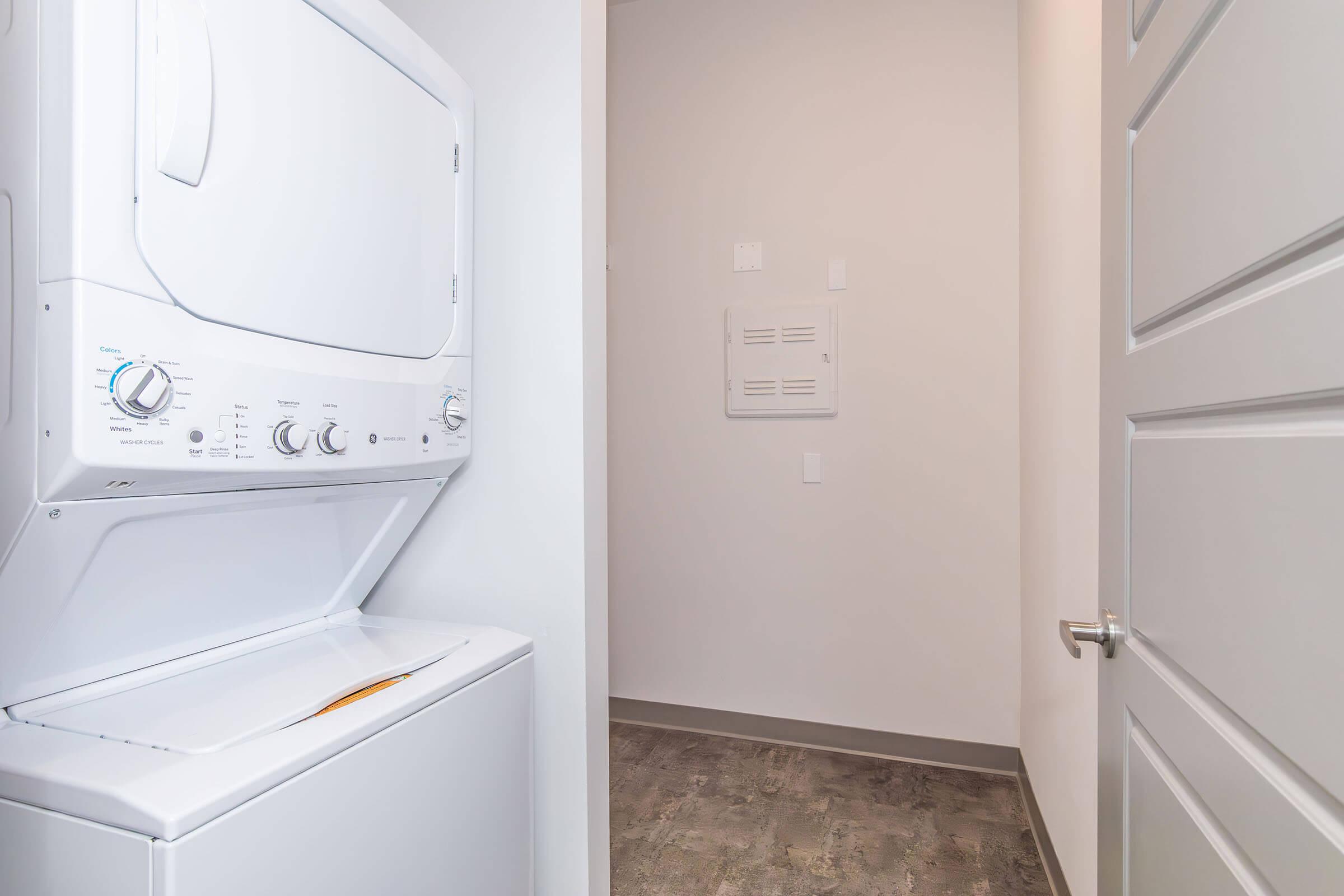 A compact laundry area featuring a stacked washer and dryer unit against a light-colored wall. The space is simple and modern, with a door leading to another room, and a utility panel visible on the wall. The floor is covered in a modern gray material.