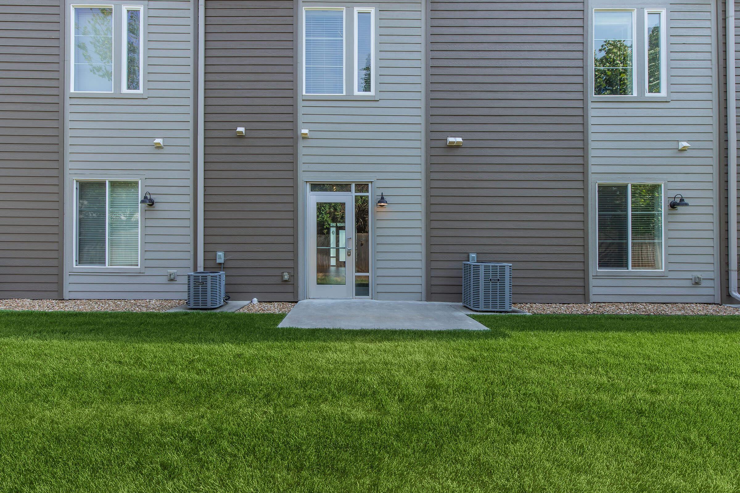 Backyard view of a modern multi-unit building featuring two air conditioning units on either side, a concrete patio leading to a glass door, and well-maintained green grass. The exterior walls are painted in shades of gray, with several windows letting in natural light.