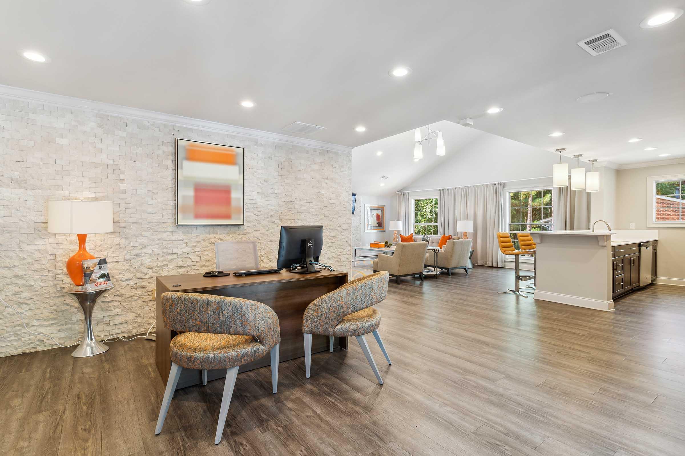 Modern interior of a welcoming lobby with a desk and computer, two stylish chairs, and a decorative lamp. The wall features a textured design, while large windows allow natural light to fill the space. Visible seating area includes cozy chairs and a kitchen bar with orange accents, creating a vibrant and inviting atmosphere.
