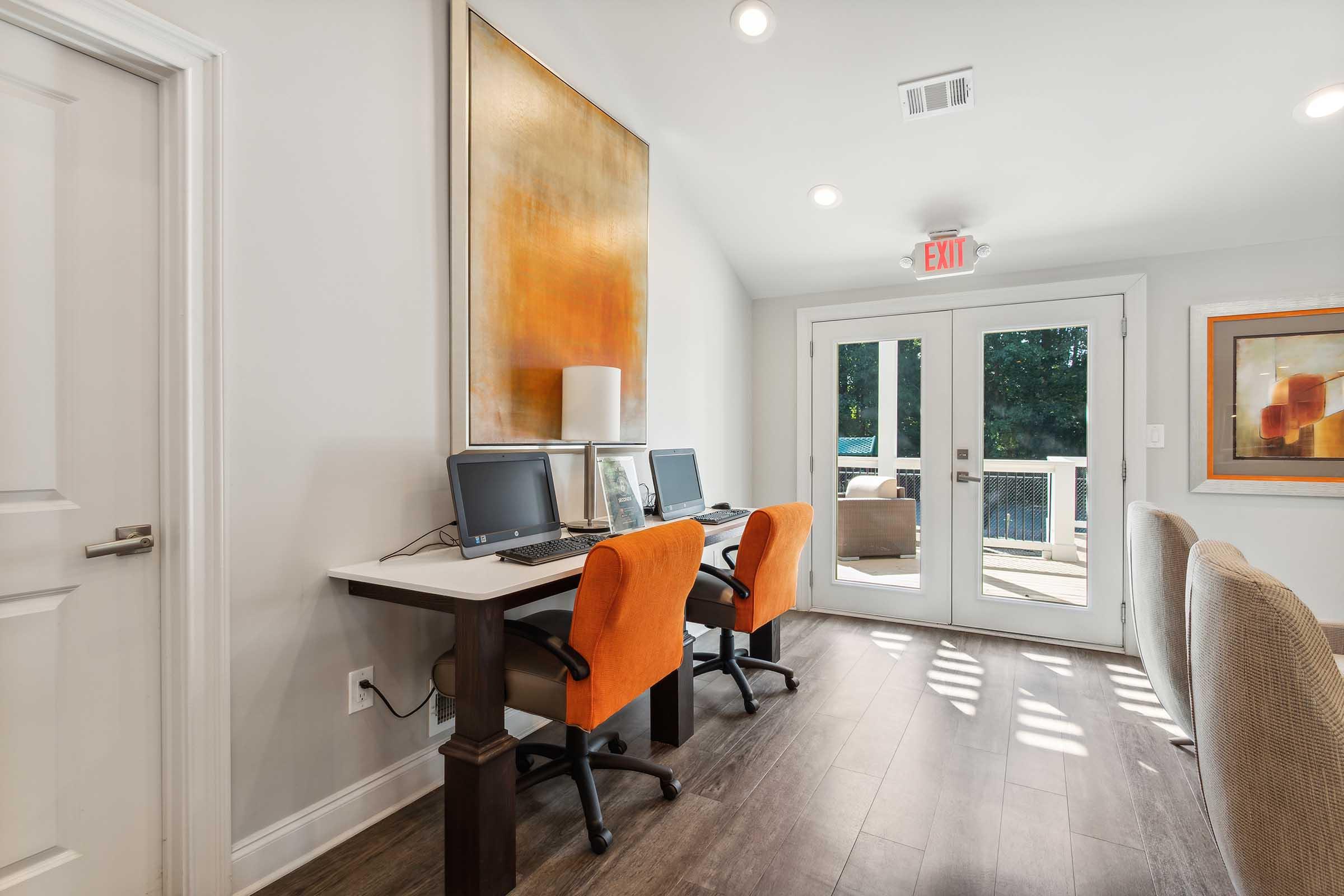 Two computer desks with orange chairs in a bright room, featuring a large abstract painting on the wall. A door leads to an outdoor area, and there are windows allowing natural light. The floor is wooden, contributing to a modern and inviting atmosphere.