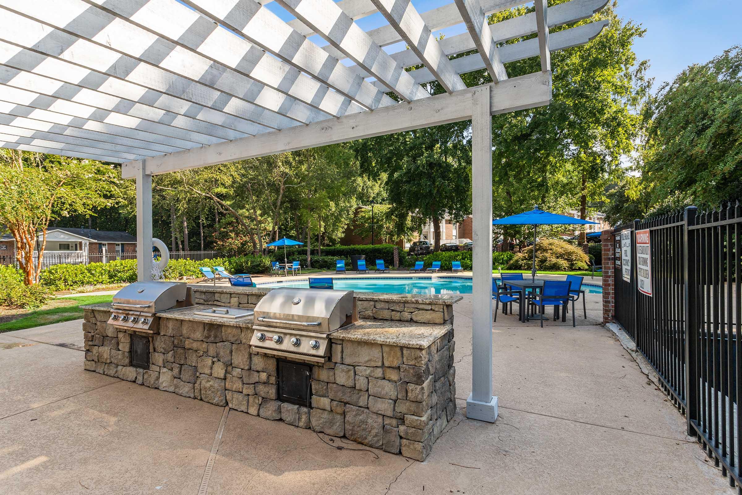Outdoor pool area featuring a stone barbecue grill station, tables with blue umbrellas, and lounge chairs surrounding a sparkling swimming pool, all set under a wooden trellis with a clear sky and lush green trees in the background.