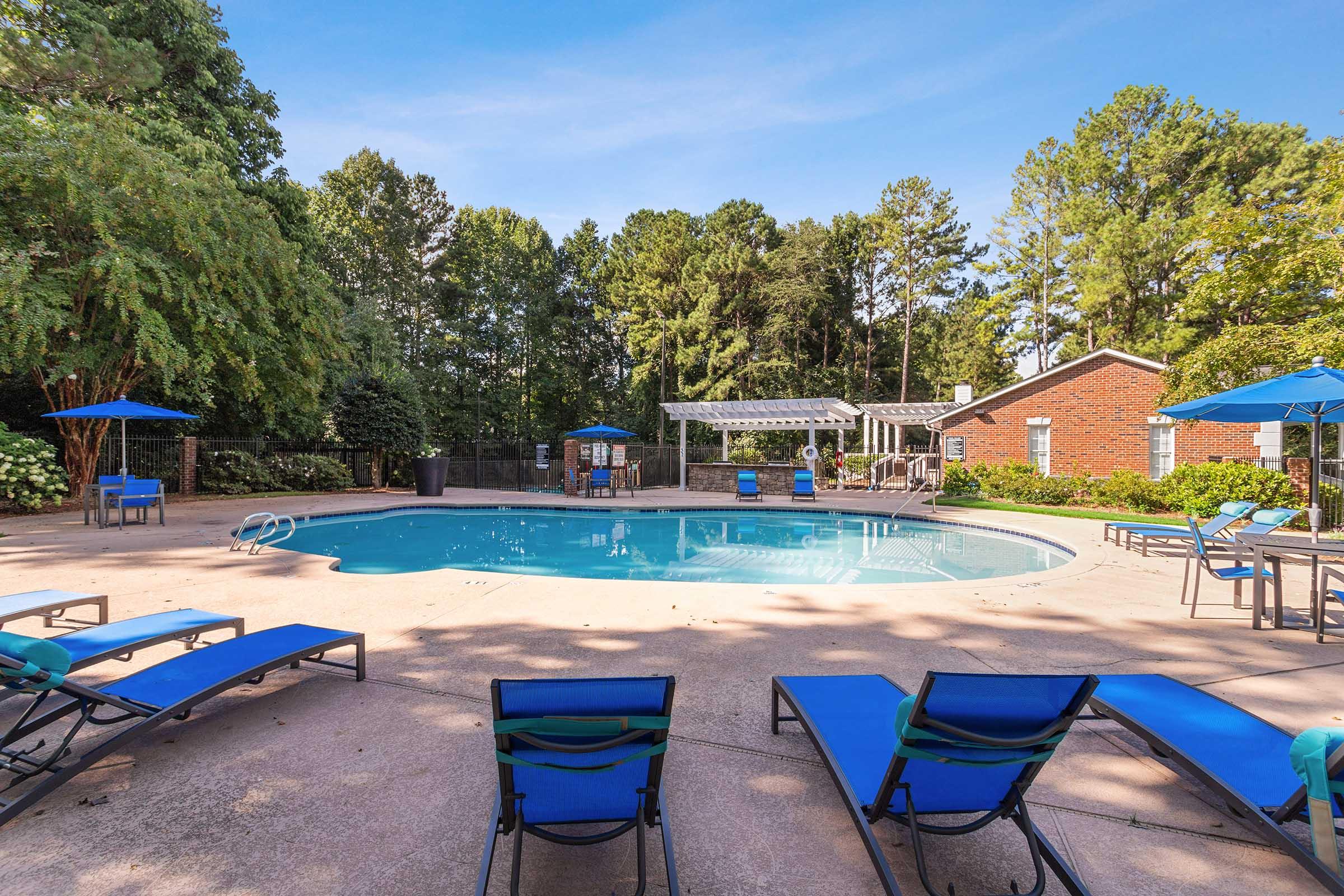 A clear blue swimming pool surrounded by lounge chairs and umbrellas, set in a lush green environment with trees. A patio area with a pergola and additional seating is visible in the background, complementing a serene outdoor space perfect for relaxation.