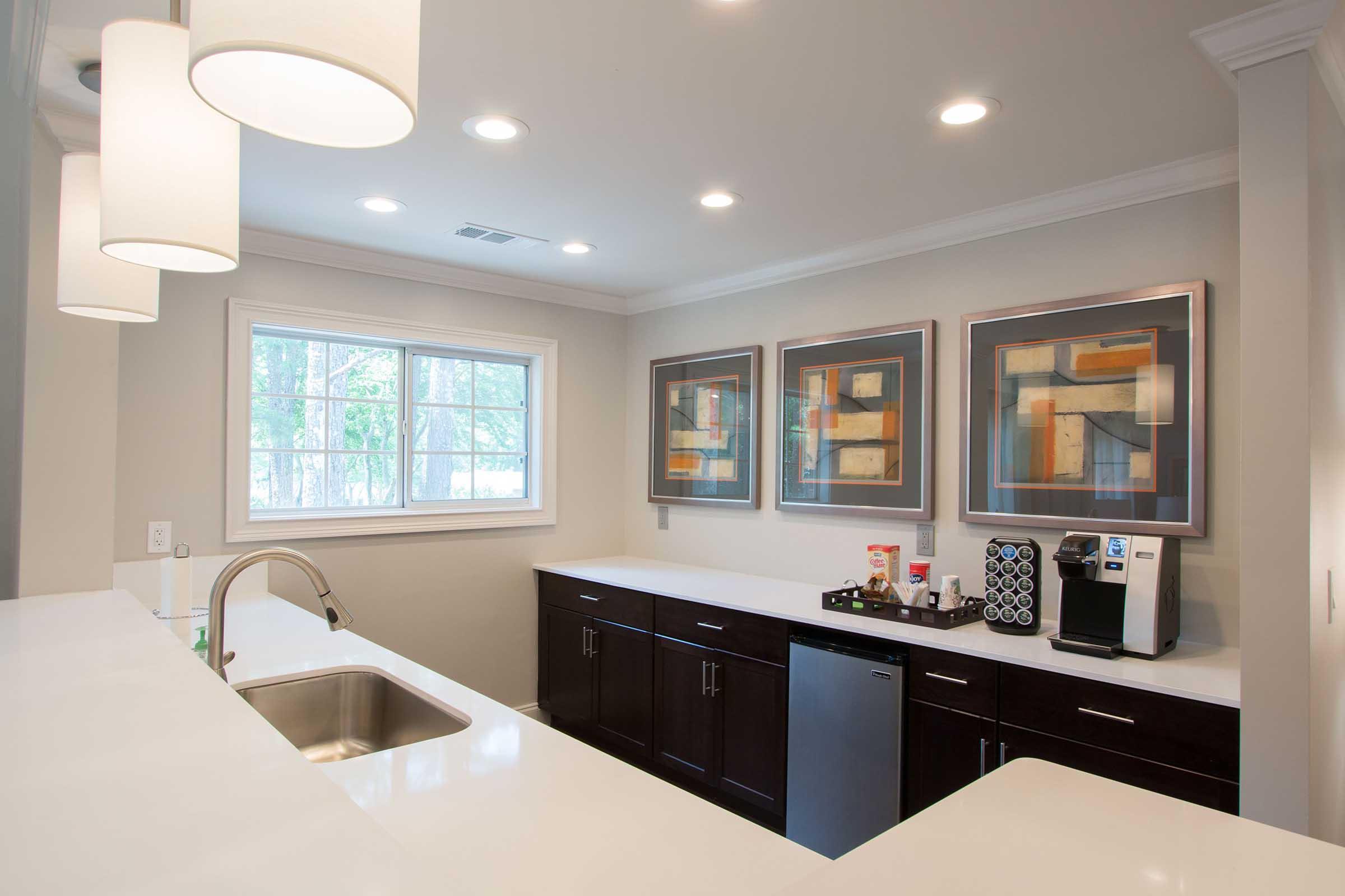 Modern kitchen with sleek dark cabinetry and a white countertop. Includes a sink, coffee maker, and a collection of artwork on the wall. Bright lighting from pendant fixtures and natural light streaming through a window. Decor features geometric designs in neutral tones.