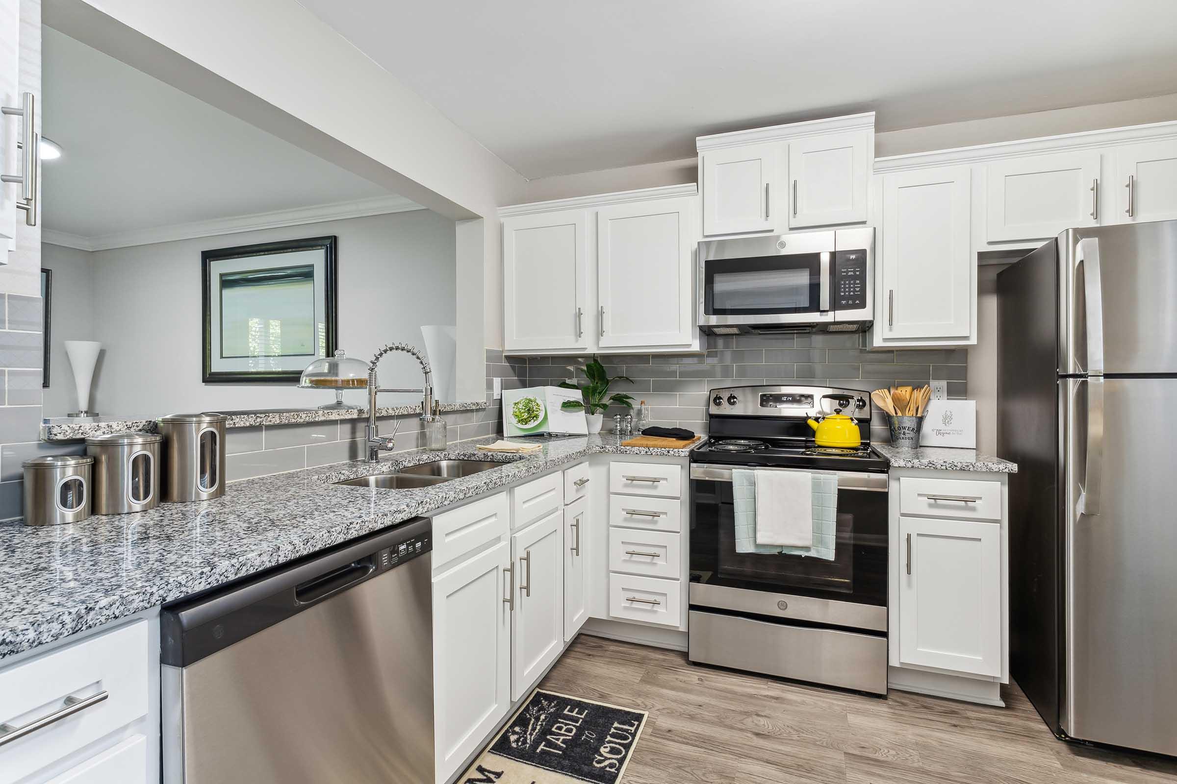 Modern kitchen featuring white cabinetry, granite countertops, stainless steel appliances, including a refrigerator, stove, and dishwasher. A small window allows natural light, and decorative items are placed on the countertops, enhancing the contemporary feel of the space.