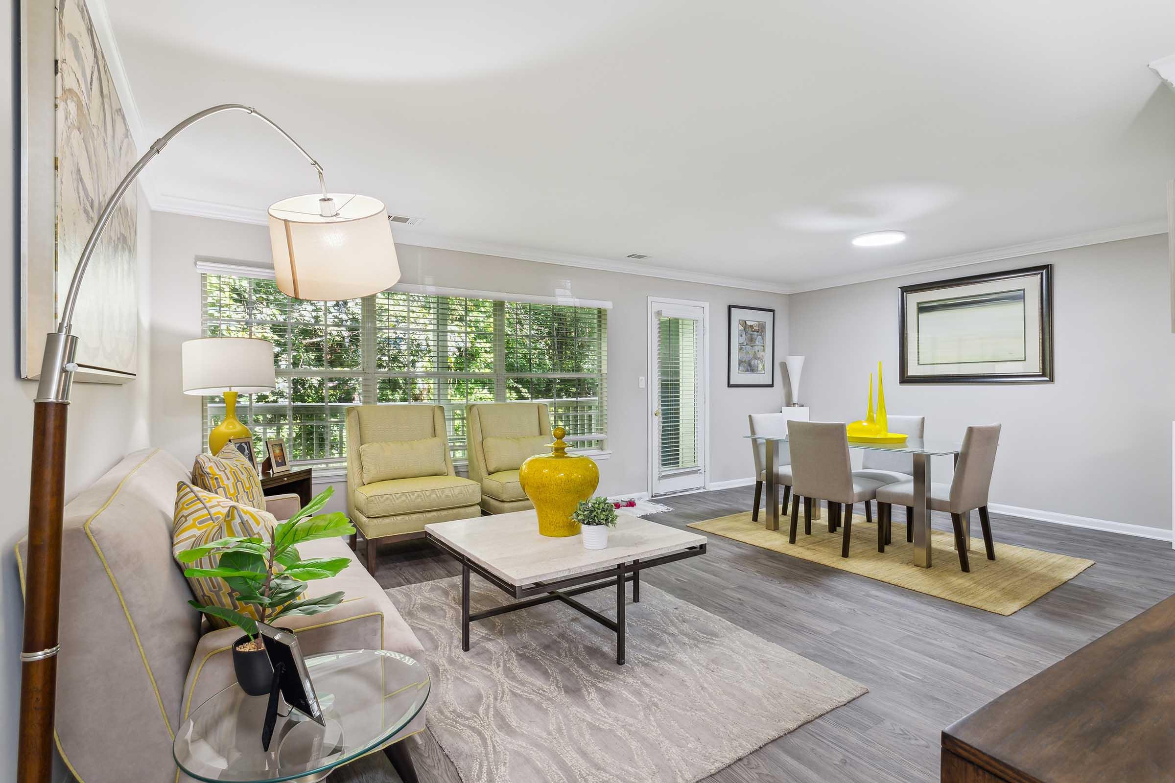A modern living room featuring a beige sofa with yellow accents, a glass coffee table with decorative plants, and a floor lamp. Adjacent is a dining area with a glass table surrounded by black chairs and a yellow centerpiece. Large windows allow natural light, complemented by neutral wall colors and wooden flooring.