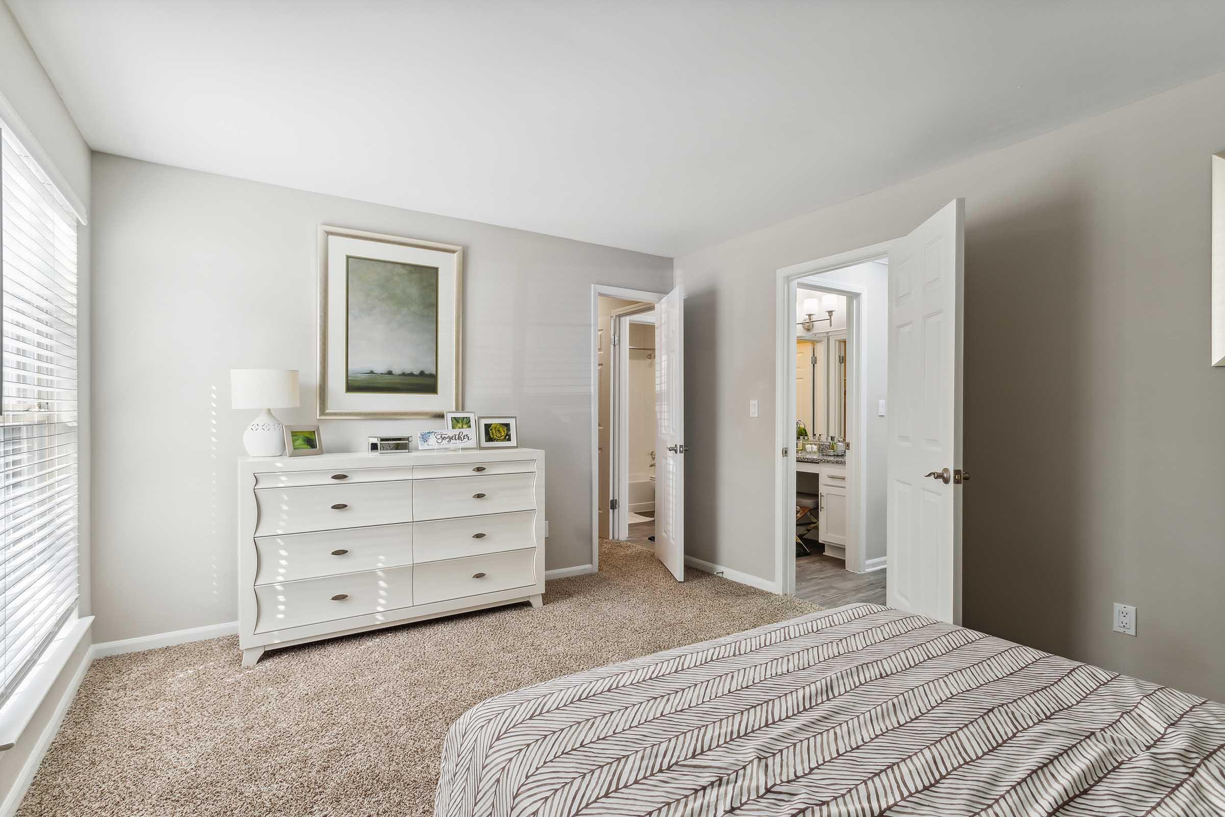A well-lit bedroom featuring a neatly made bed with a striped comforter. A white dresser with decorative items is positioned against a light gray wall, and a doorway leads to a bathroom. The room has large windows with blinds, allowing natural light to brighten the space.