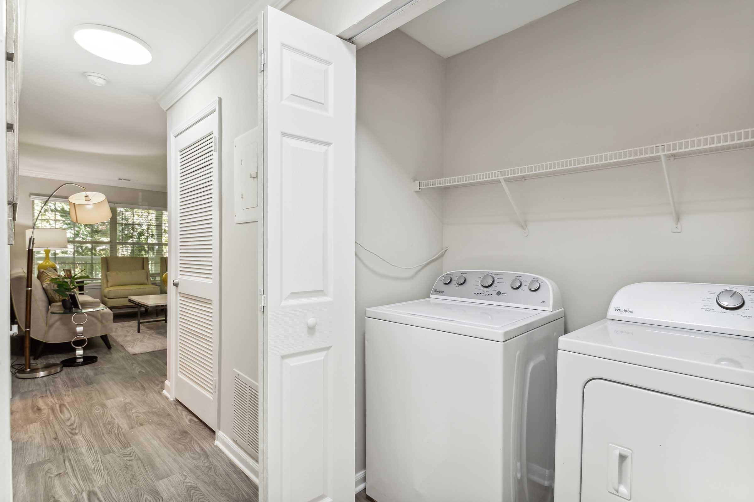 A spacious laundry room featuring a washer and dryer side by side, with a shelf above for storage. The room has light-colored walls and a doorway leading to a bright living area with natural light. A comfortable seating area can be seen in the background, creating an inviting space.