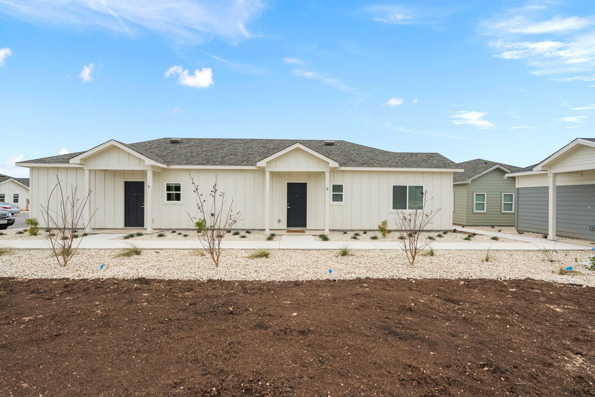 A modern duplex home with a light-colored façade and black doors, set against a clear blue sky. The front yard features landscaped areas with gravel and small plants, while the surrounding neighborhood includes additional homes visible in the background.