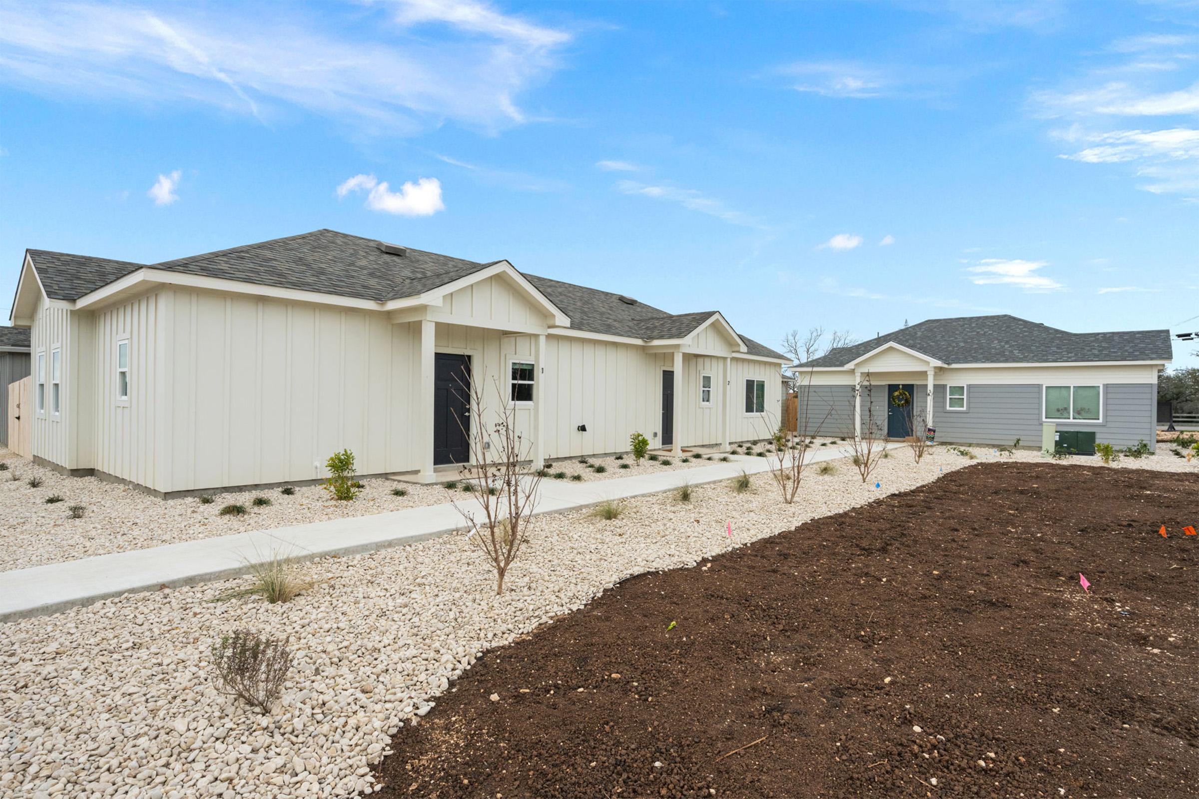 Two single-story houses with modern designs, one featuring a light-colored exterior and the other a gray facade. A concrete pathway runs between them, surrounded by gravel landscaping with sparse plants. The sky is clear and bright, indicating a sunny day.