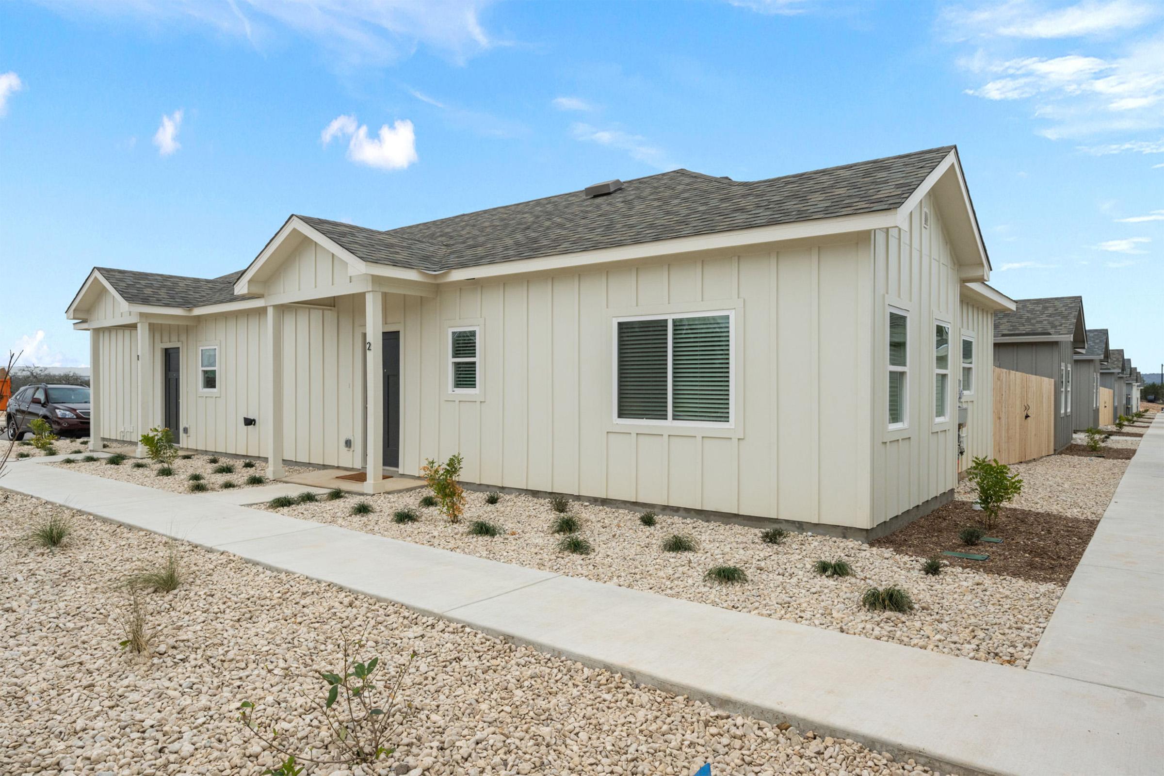A modern single-story house with a light-colored exterior and a sloped roof, situated in a neighborhood. The front features a concrete walkway and landscaped areas with small plants and rocks. There are several similar houses in the background, set against a clear blue sky.