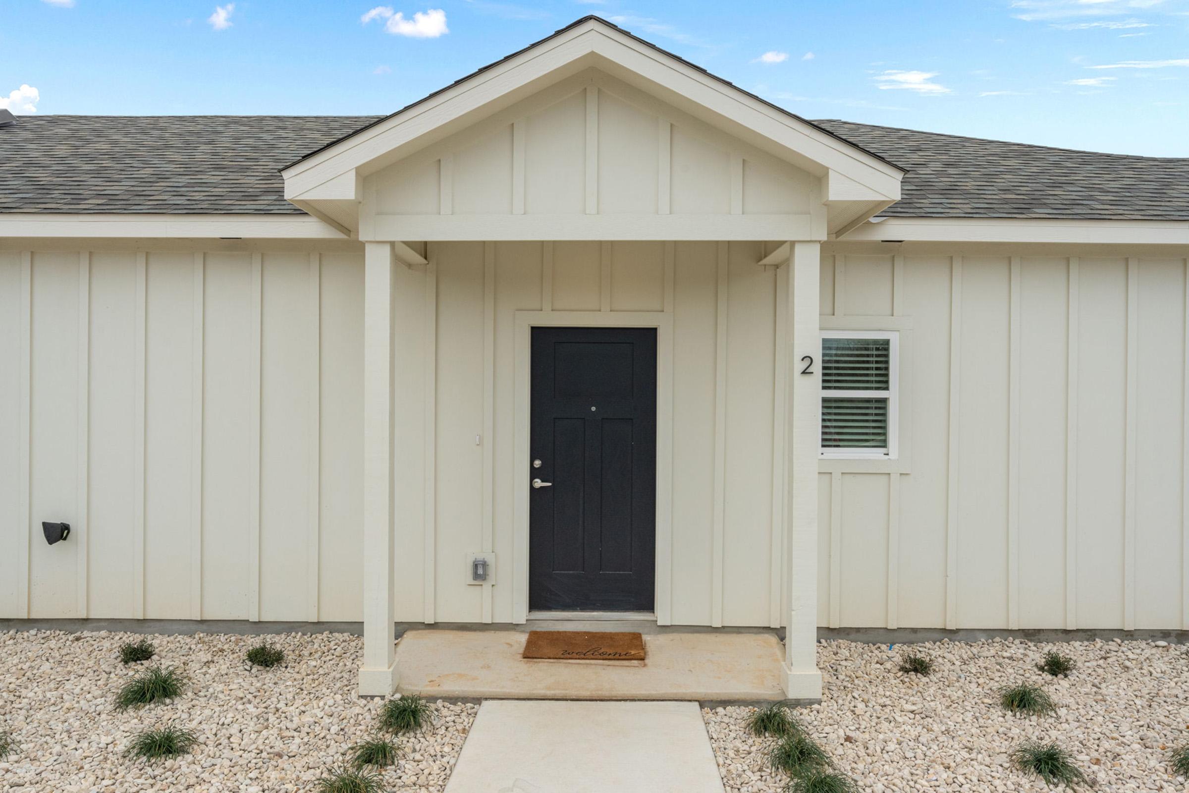 A modern, single-story house entryway featuring a dark front door with a small porch, framed by a gabled roof. The exterior is painted in light beige with horizontal paneling. A welcome mat is placed at the entrance, and the area is landscaped with small rocks and sparse grass.