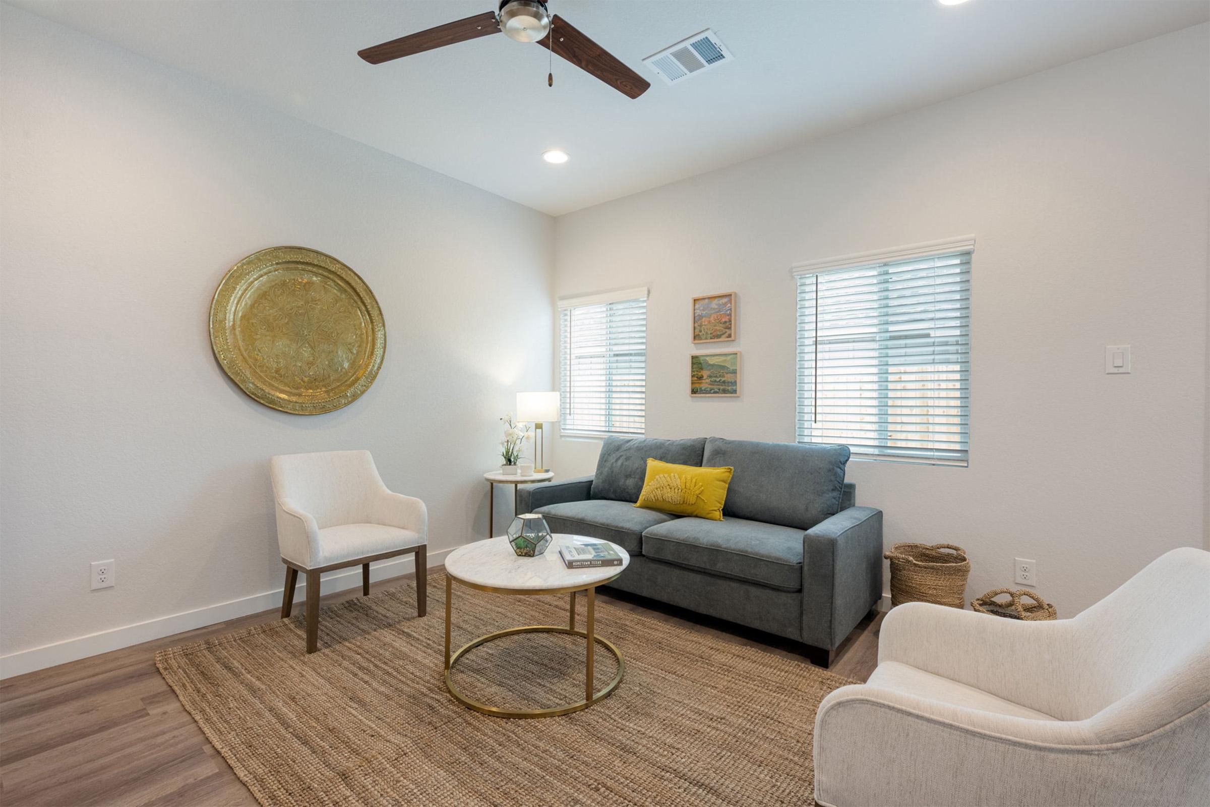 A cozy living room featuring a gray sofa with yellow cushions, two white armchairs, a round coffee table, and a textured area rug. Natural light fills the space through window blinds, while a decorative wall plate and small plants add a touch of decor.