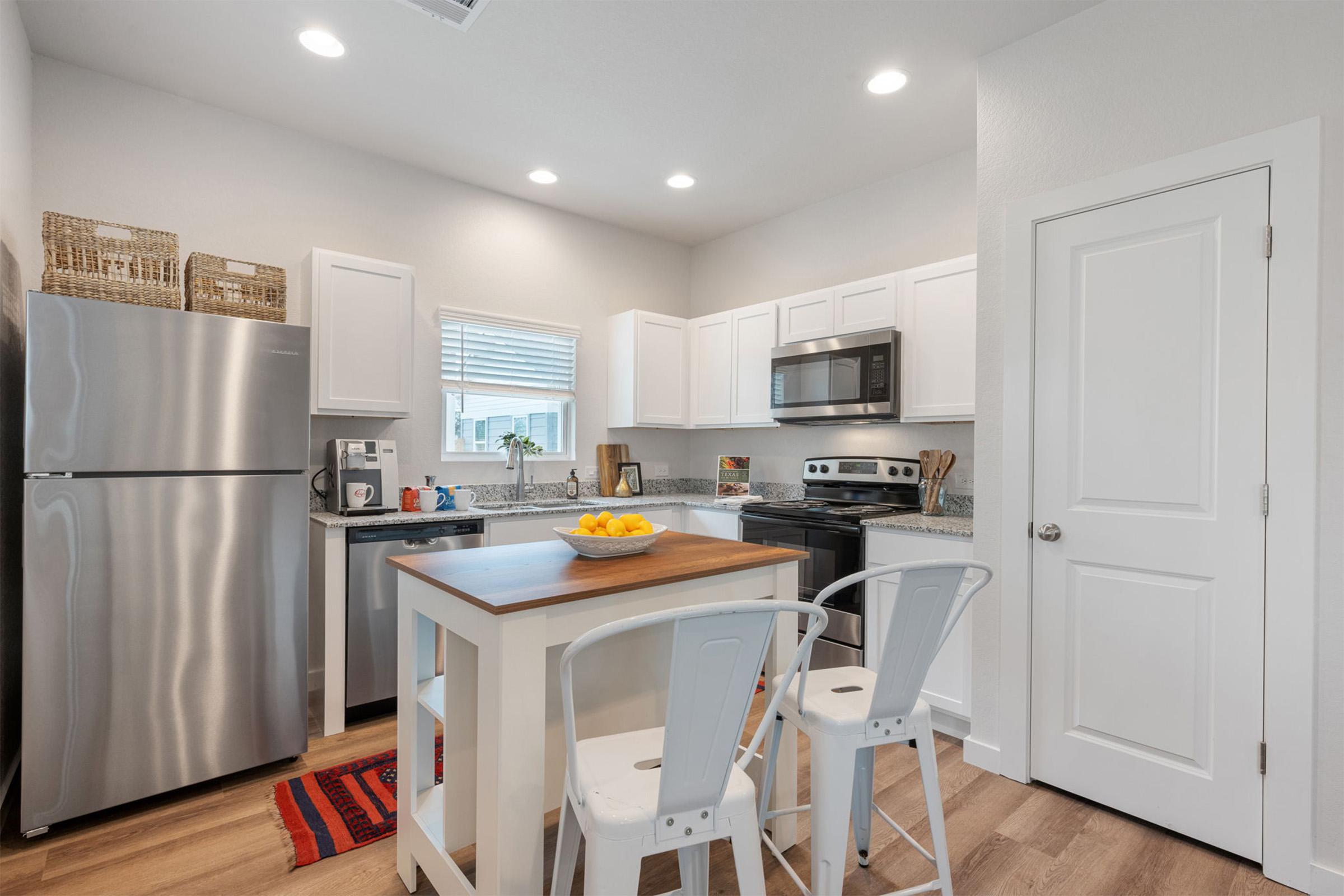 Modern kitchen featuring white cabinets, stainless steel appliances including a refrigerator and microwave, and a wood island with two white bar stools. The countertop has a bowl of fruit and there are decorative baskets on shelves. The space is well-lit with overhead lights and has a warm, inviting feel.
