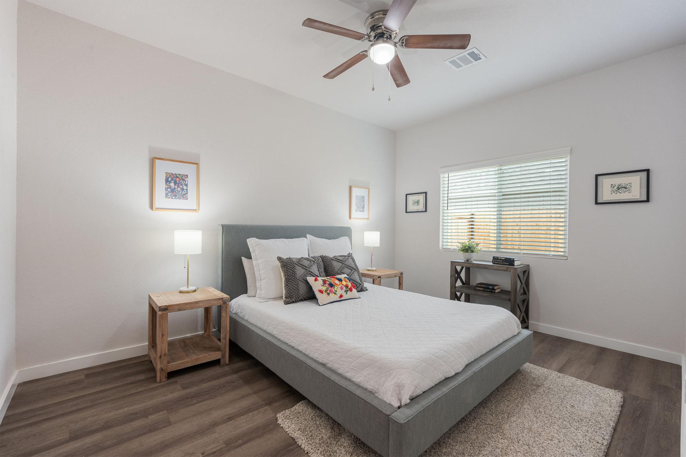 A modern, airy bedroom featuring a light gray bed with white bedding, wooden nightstands with lamps on either side, and artwork on the walls. A window lets in natural light, and a small shelf holds decorative items. The floor is covered with a plush rug, creating a cozy atmosphere.