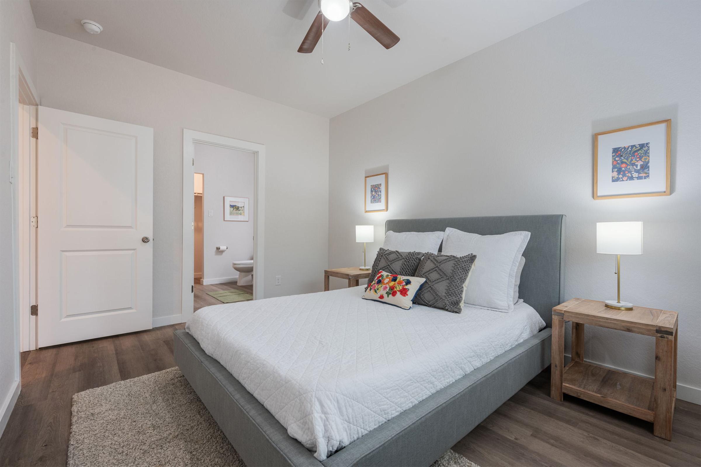 A well-lit bedroom featuring a gray upholstered bed with white bedding, two wooden nightstands with lamps, and colorful framed artwork on the walls. A door leads to a bathroom visible in the background. The room has a light-colored, modern design with wood flooring.