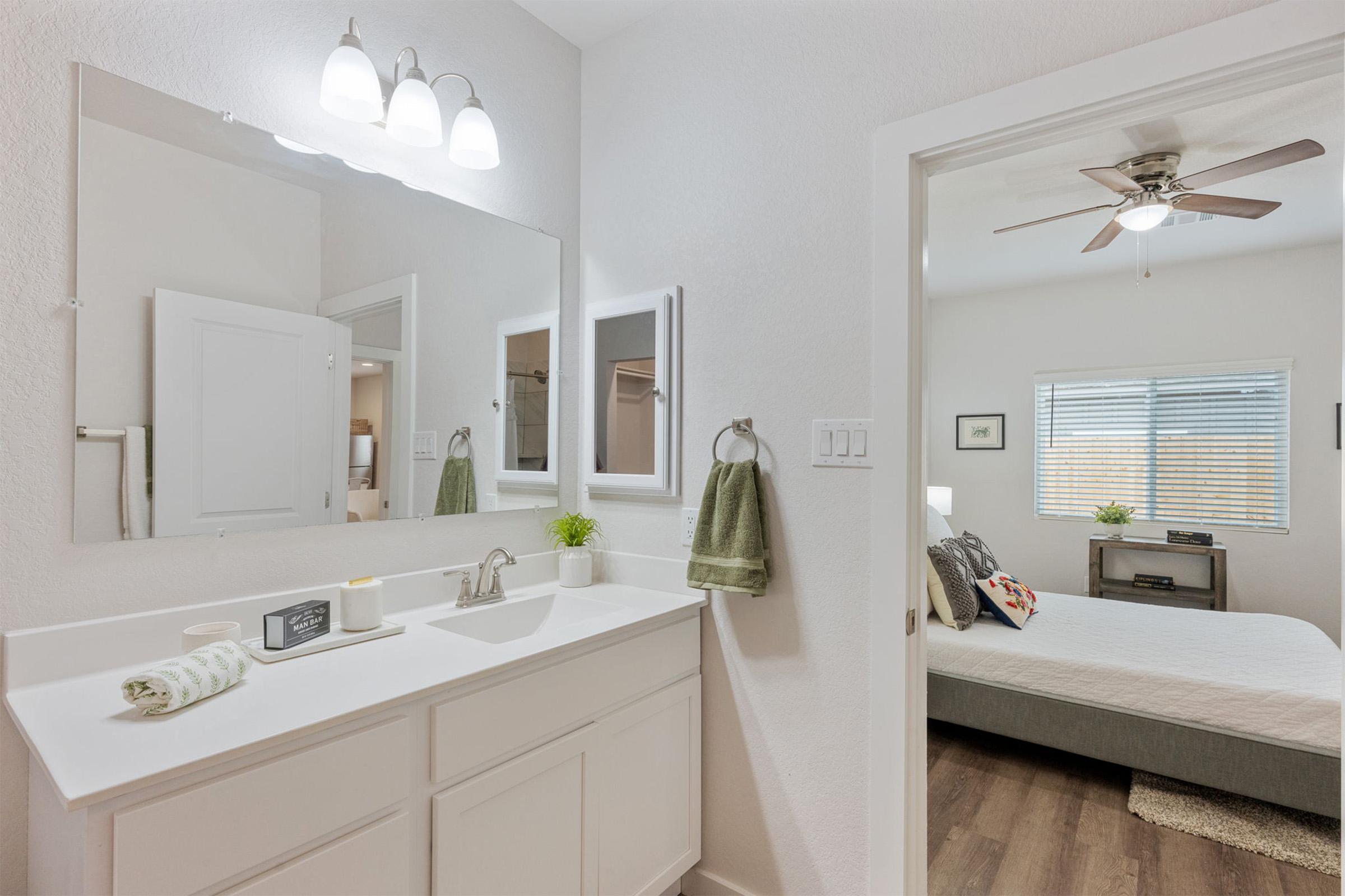 A bright bathroom featuring a white vanity with a sink, a large mirror, and modern light fixtures. There's a neatly folded towel hanging on the side, and a small plant on the countertop. The doorway leads to a bedroom with a bed, ceiling fan, and window, creating a spacious and inviting atmosphere.