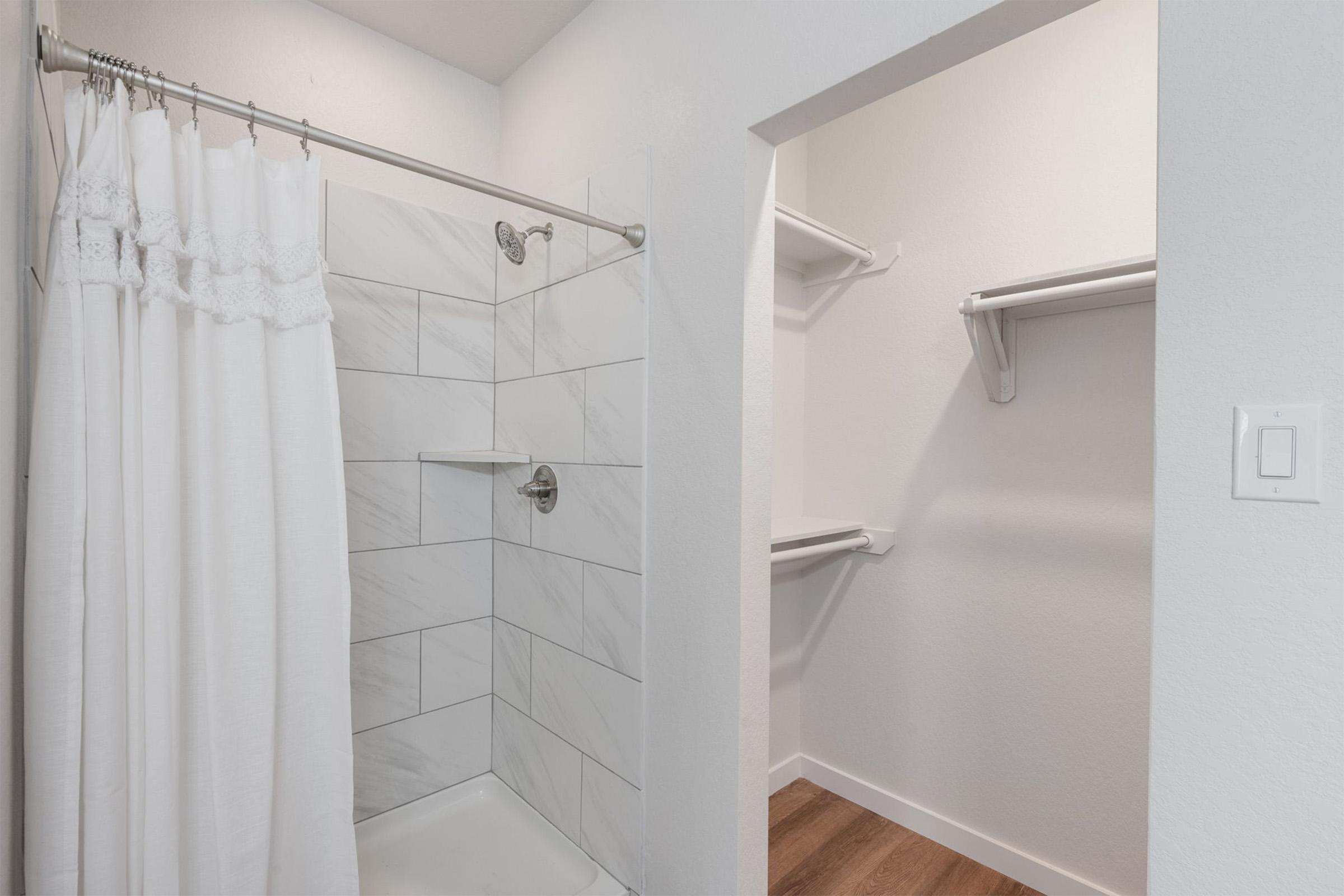 A modern bathroom featuring a white shower curtain hanging over a tiled shower with a sleek showerhead. To the right, there is a small walk-in closet with blank walls and two shelves. The flooring is a warm wood tone, creating a cozy atmosphere.