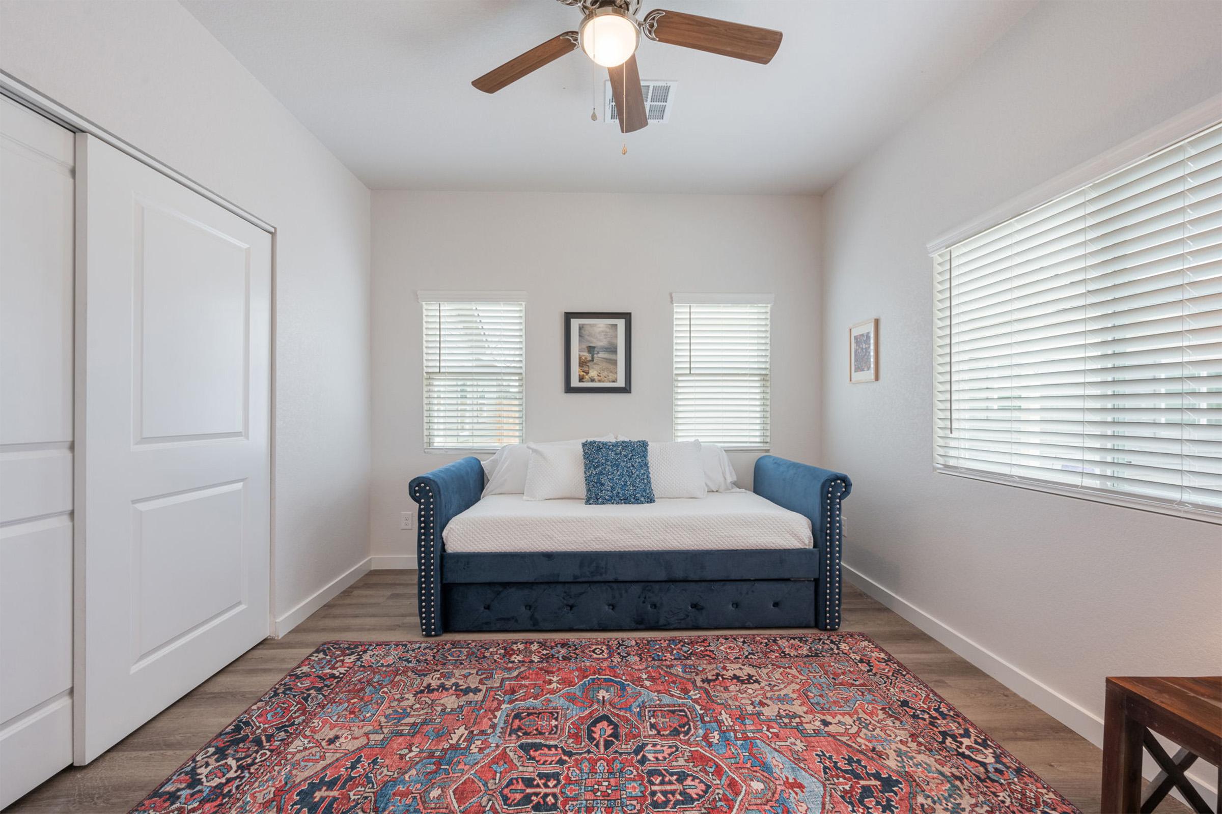 A cozy, well-lit room featuring a blue daybed with white bedding, a decorative blue pillow, and an intricate area rug. The walls are painted in a light color, complemented by two windows with blinds, a framed picture on the wall, and a ceiling fan, creating a warm and inviting atmosphere.