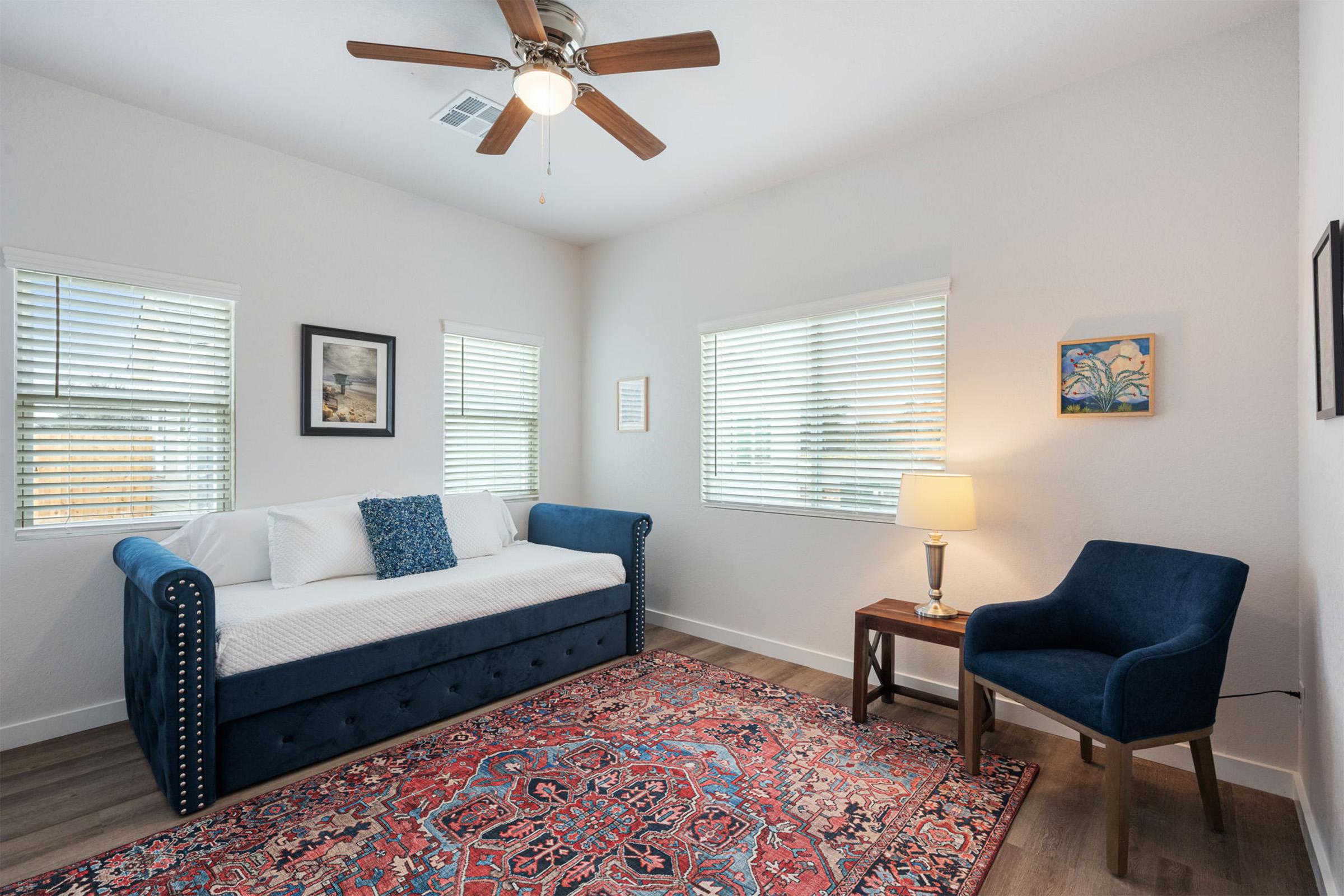 A cozy, well-lit room featuring a blue daybed with white cushions, a dark blue armchair, a wooden side table with a lamp, and decorative wall art. Light-colored walls and two windows with blinds allow natural light to fill the space. A colorful area rug adds warmth to the modern decor.