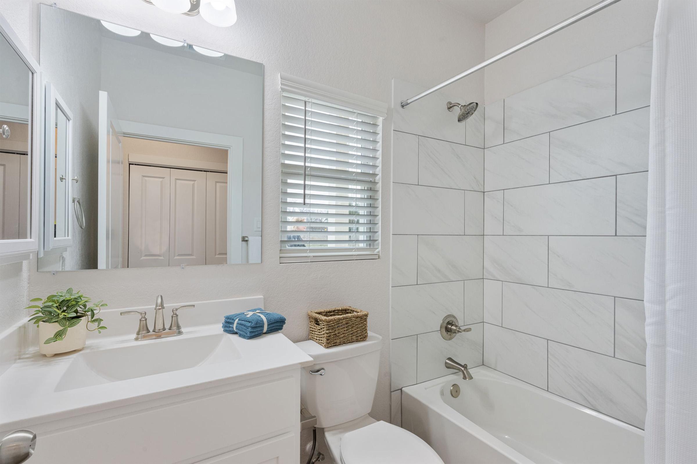 A clean and modern bathroom featuring a white bathtub and shower combination. The sink area includes a mirror, faucet, and a small potted plant. Several blue towels are neatly arranged on the countertop, and a wicker basket adds storage. Natural light filters through a window with blinds.