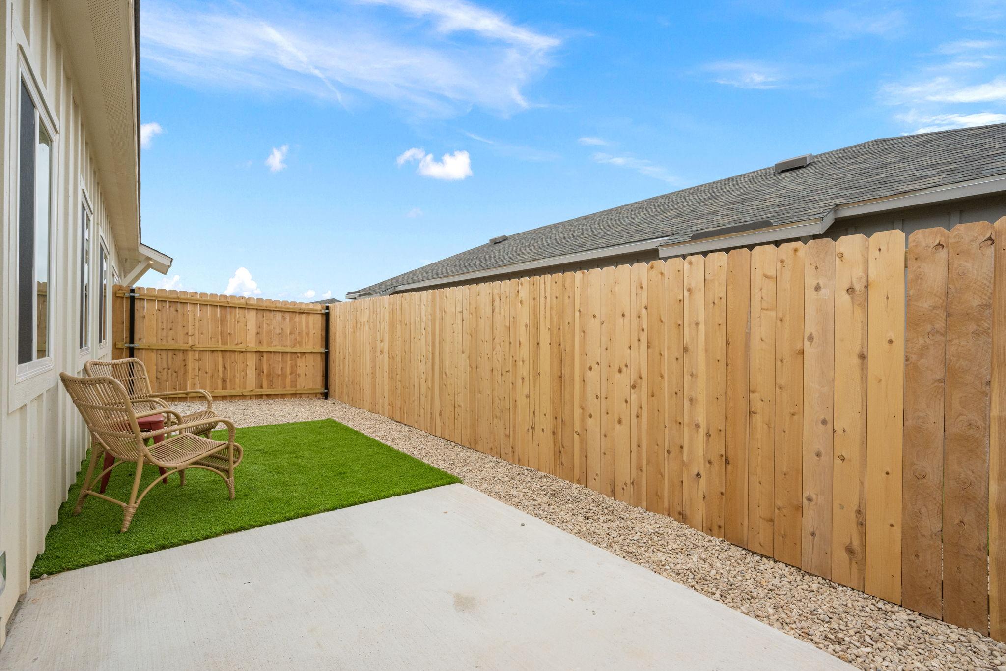 A narrow outdoor space featuring a light-colored concrete patio, bordered by wooden fencing. There are two rattan chairs on the left side and a patch of artificial grass on the right, with gravel filling the rest of the area. The sky above is clear with a few scattered clouds.