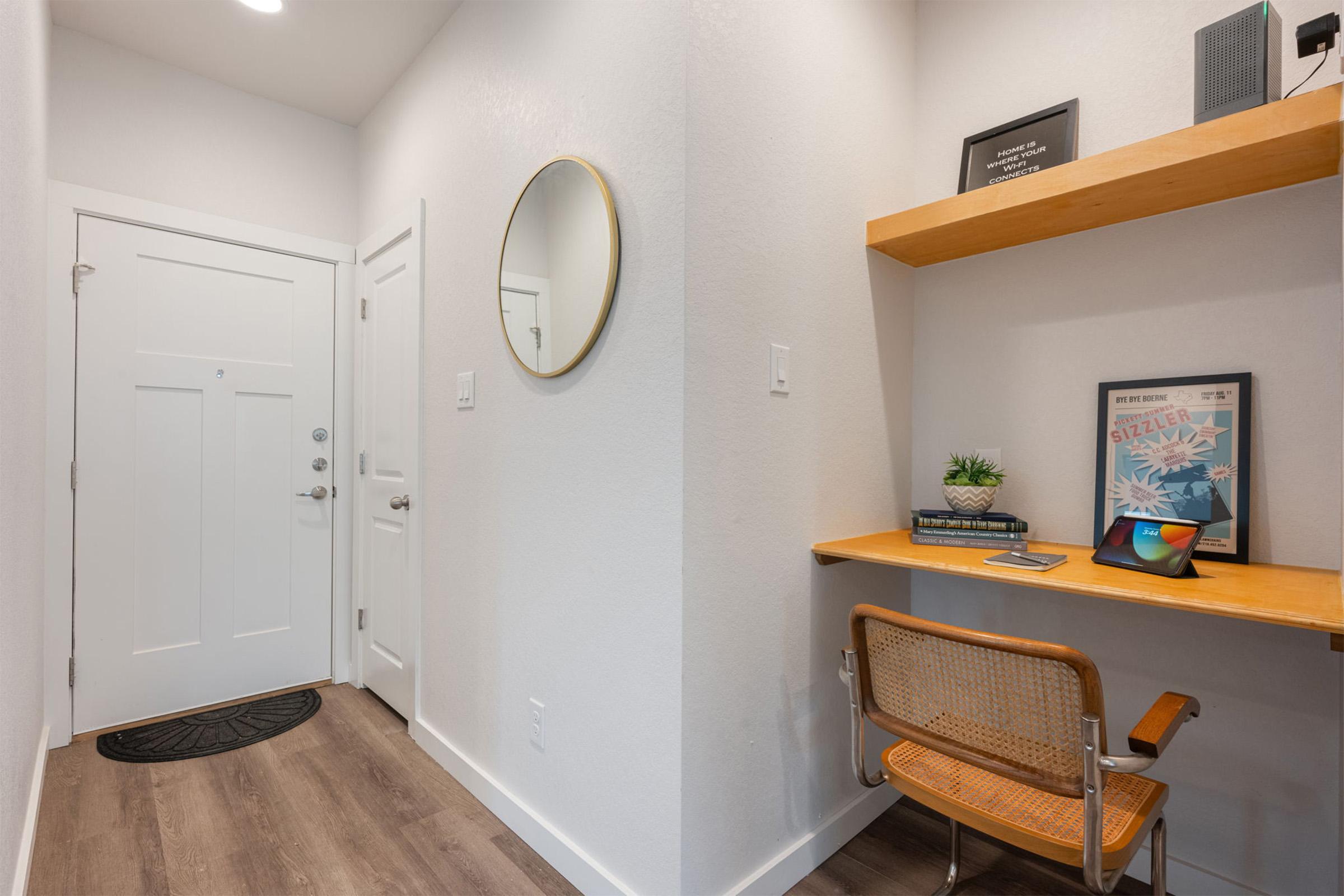 A modern entryway featuring a white door, a round mirror on the wall, and a small wooden shelf with framed artwork and a potted plant. Below the shelf, there's a desk with a chair and a tablet, along with a decorative welcome mat on the floor. The walls are painted light, creating a bright atmosphere.