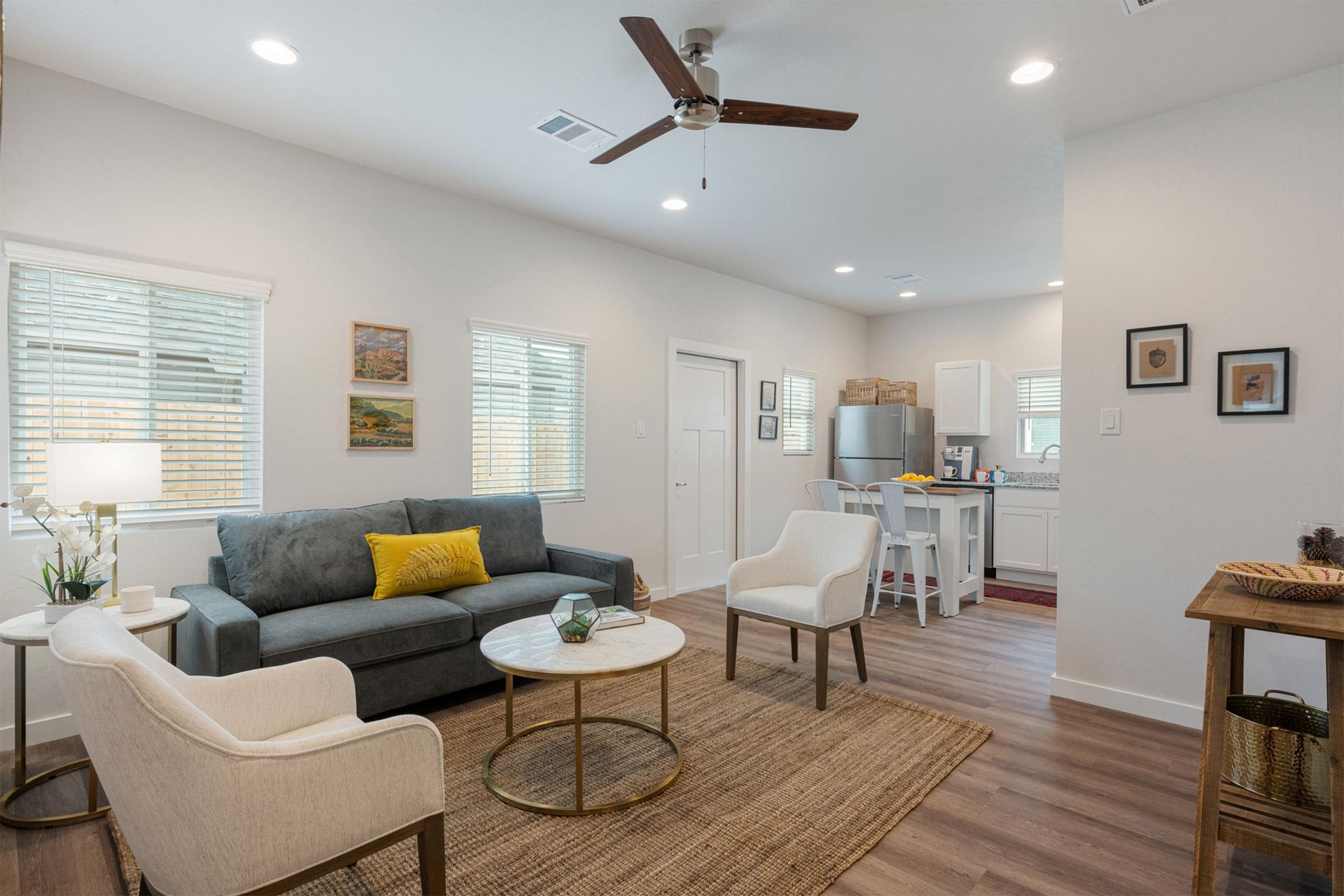 A cozy living room featuring a grey sofa with a yellow pillow, two white chairs, a circular coffee table, and a woven rug. Natural light pours in through window panes, and a small dining area with kitchen appliances is visible in the background, creating a warm and inviting atmosphere.