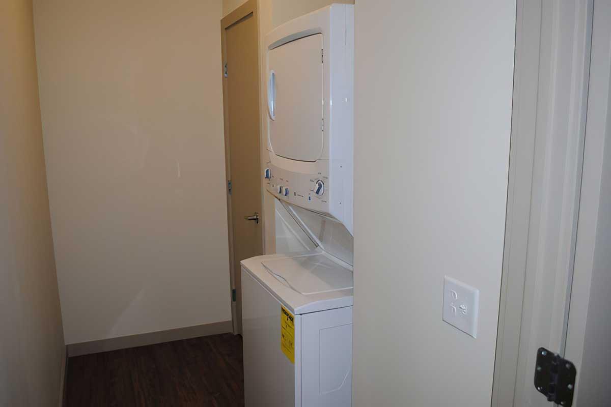 A compact laundry area featuring a stacked washer and dryer, located in a narrow hallway with beige walls and wooden flooring