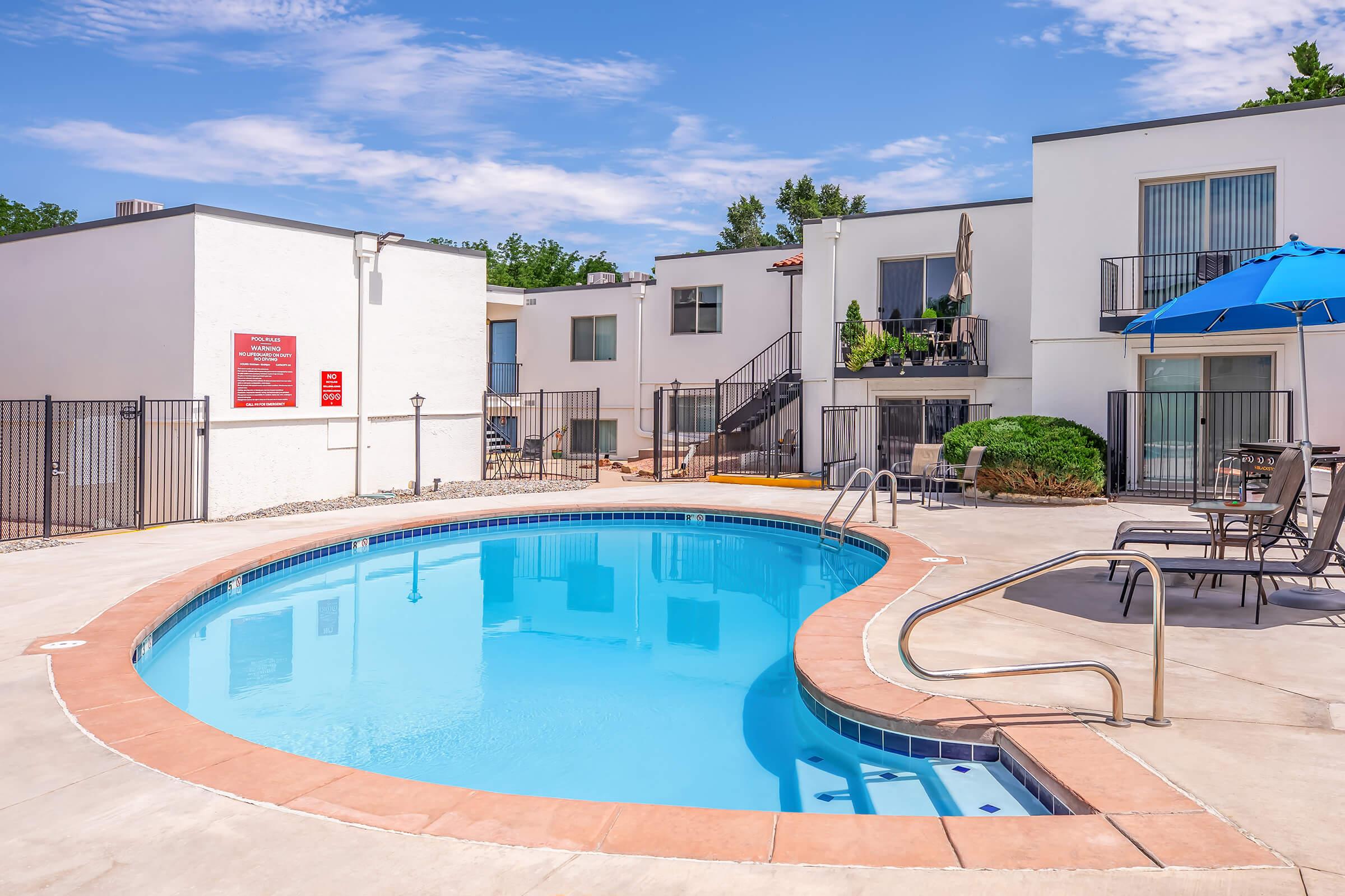 A clear blue swimming pool surrounded by a patio area and lounge chairs. White buildings with balconies are in the background, and a blue umbrella provides shade near the pool. Lush greenery adds to the relaxing atmosphere on a sunny day.