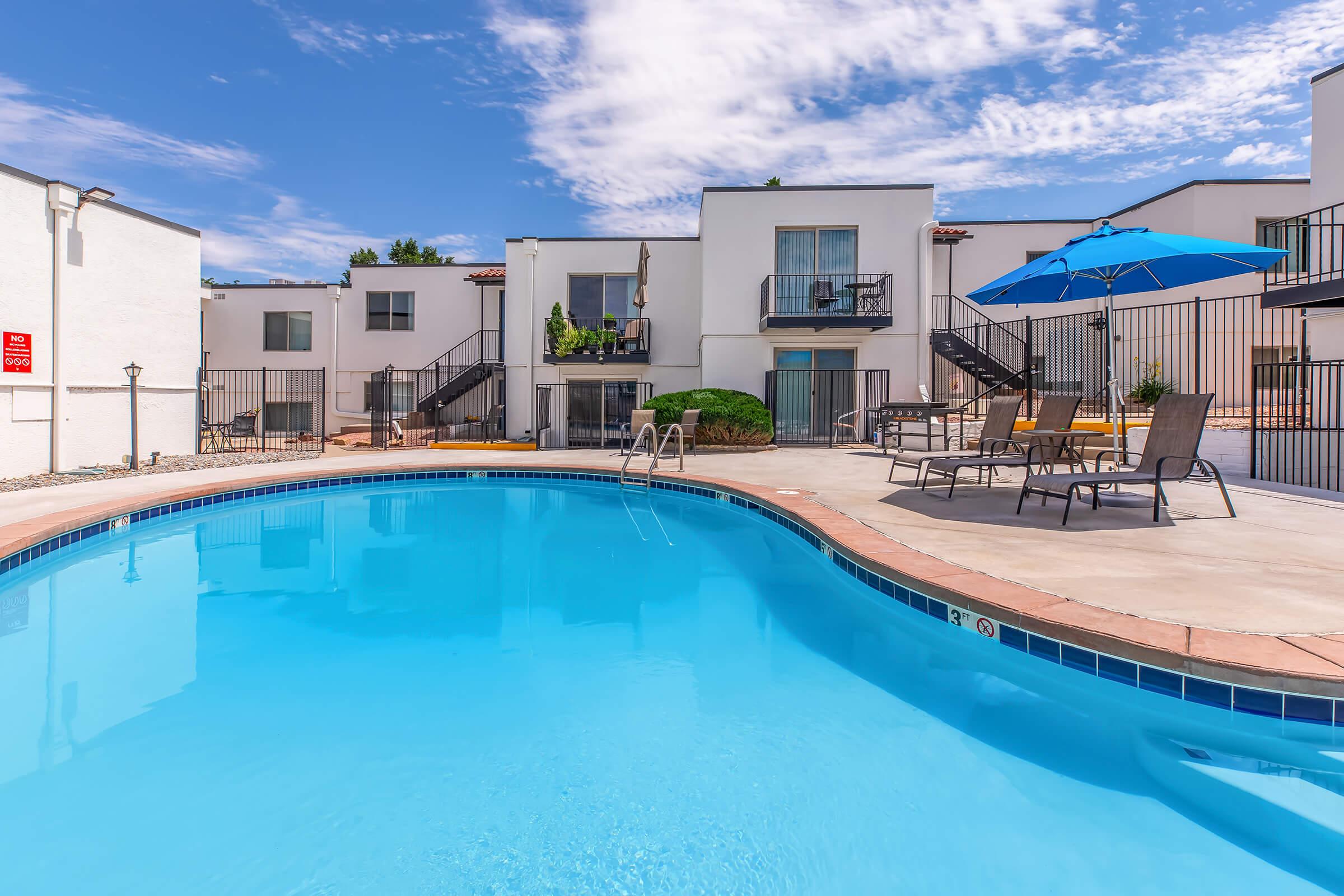 A swimming pool surrounded by lounge chairs and umbrellas, with white buildings in the background. The sky is partly cloudy, creating a bright and inviting atmosphere.