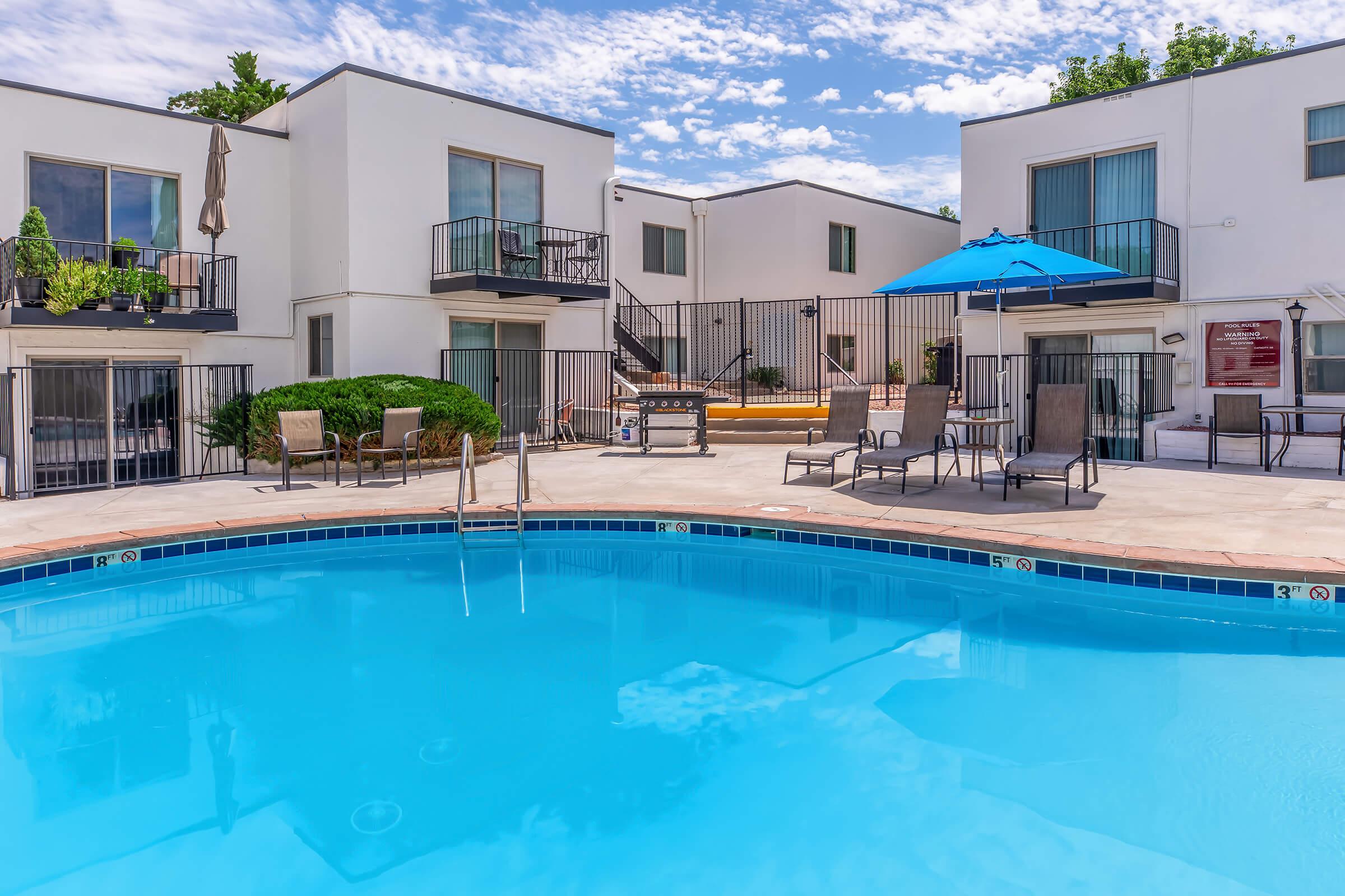 A tranquil pool area surrounded by white apartment buildings, featuring lounge chairs, an umbrella, and potted plants. The clear blue water reflects a sunny sky with some clouds, creating a relaxing outdoor atmosphere. A gated hot tub is visible in the background.