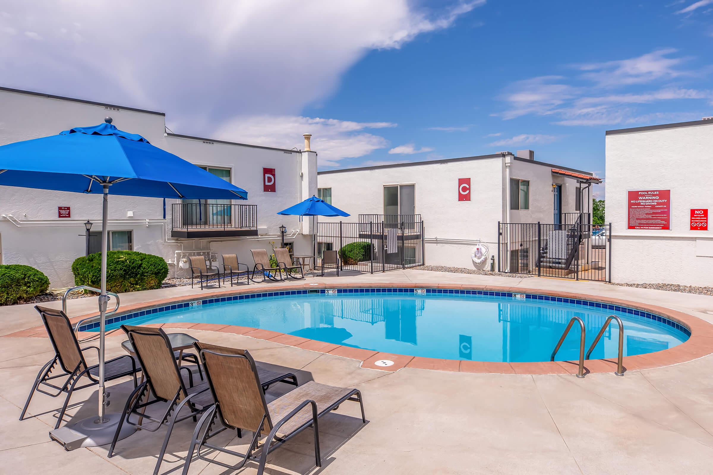 A bright outdoor pool area surrounded by lounge chairs and umbrellas. Nearby buildings are white with red accents, and there are potted plants and a safety ring near the pool's edge. The sky is partly cloudy, creating a pleasant atmosphere for relaxation.