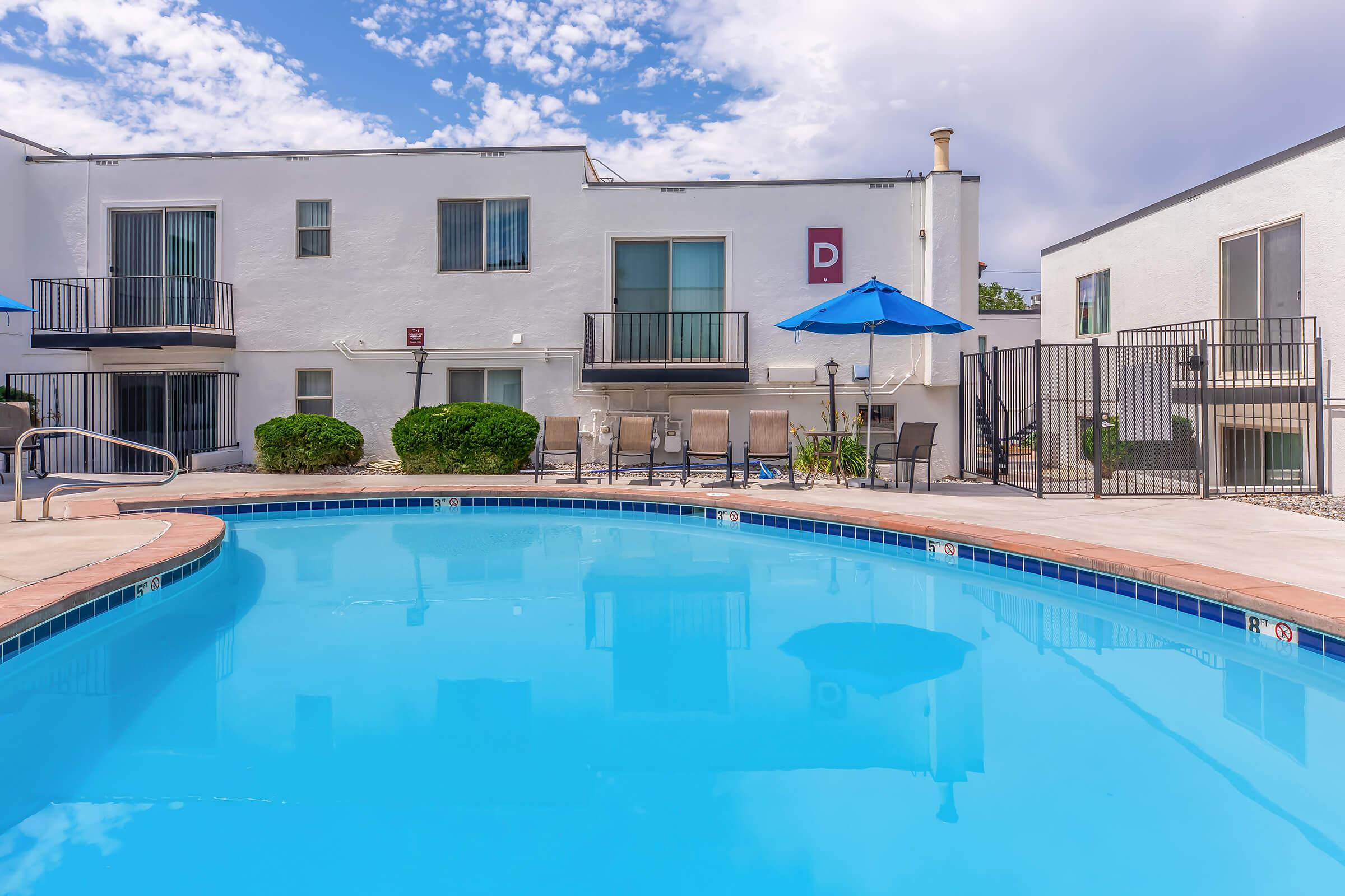 A clear blue swimming pool surrounded by lounge chairs and an umbrella, with a white building in the background. The pool area features a fence and landscaped greenery, under a partly cloudy sky. The scene conveys a relaxing and inviting atmosphere.