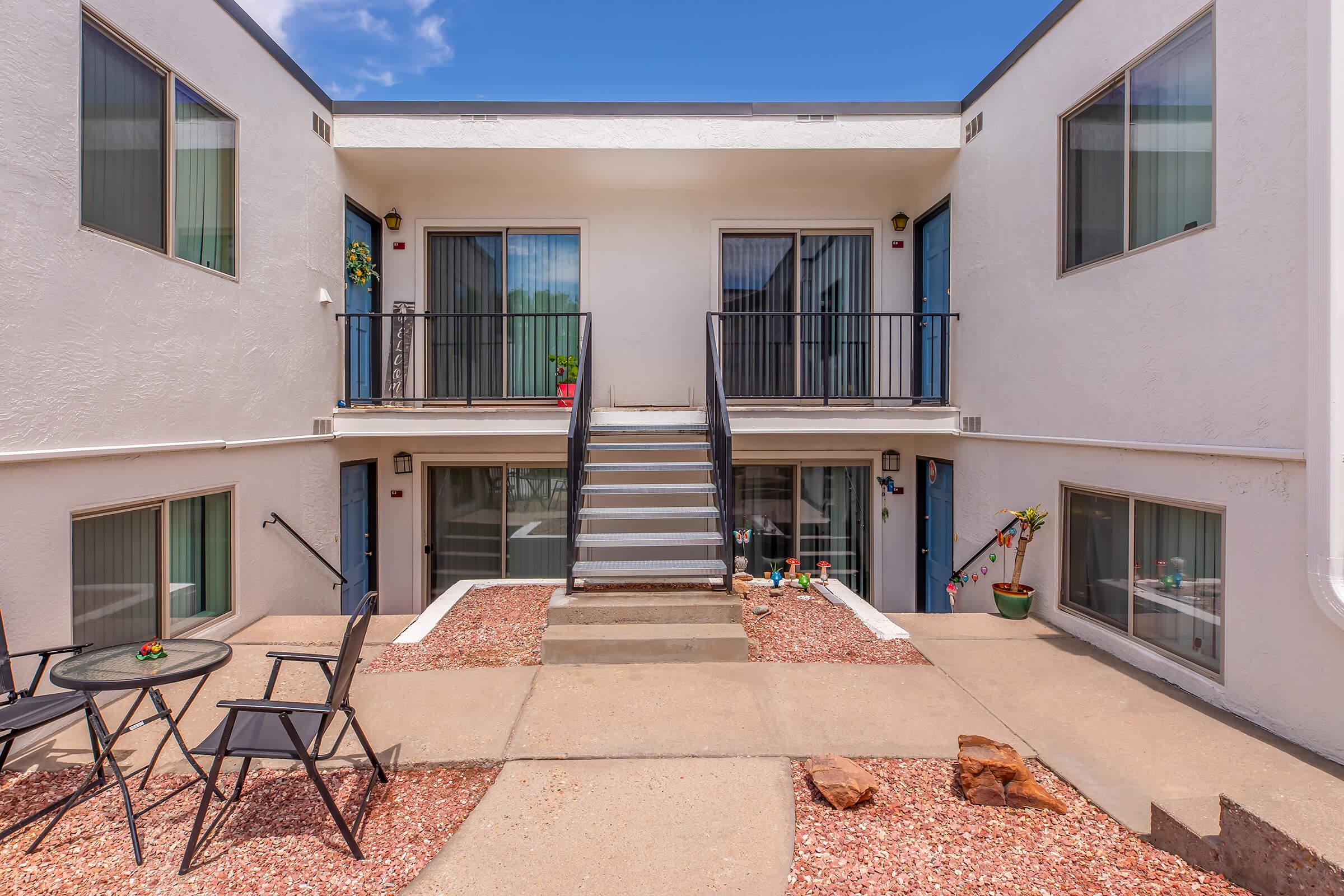 A view of a two-story apartment complex with a central staircase. The courtyard area features a concrete pathway, decorative rocks, and potted plants. Two black chairs and a small table are set up on one side, with sliding glass doors visible on both floors. Blue sky and scattered clouds are in the background.